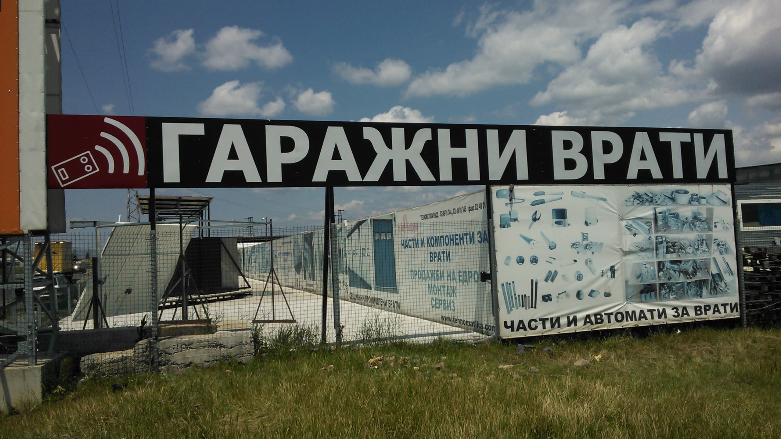 Две големи информационни пана на метална конструкция, поставени на открито пред ограда на имот, на фона на небе с облаци.
