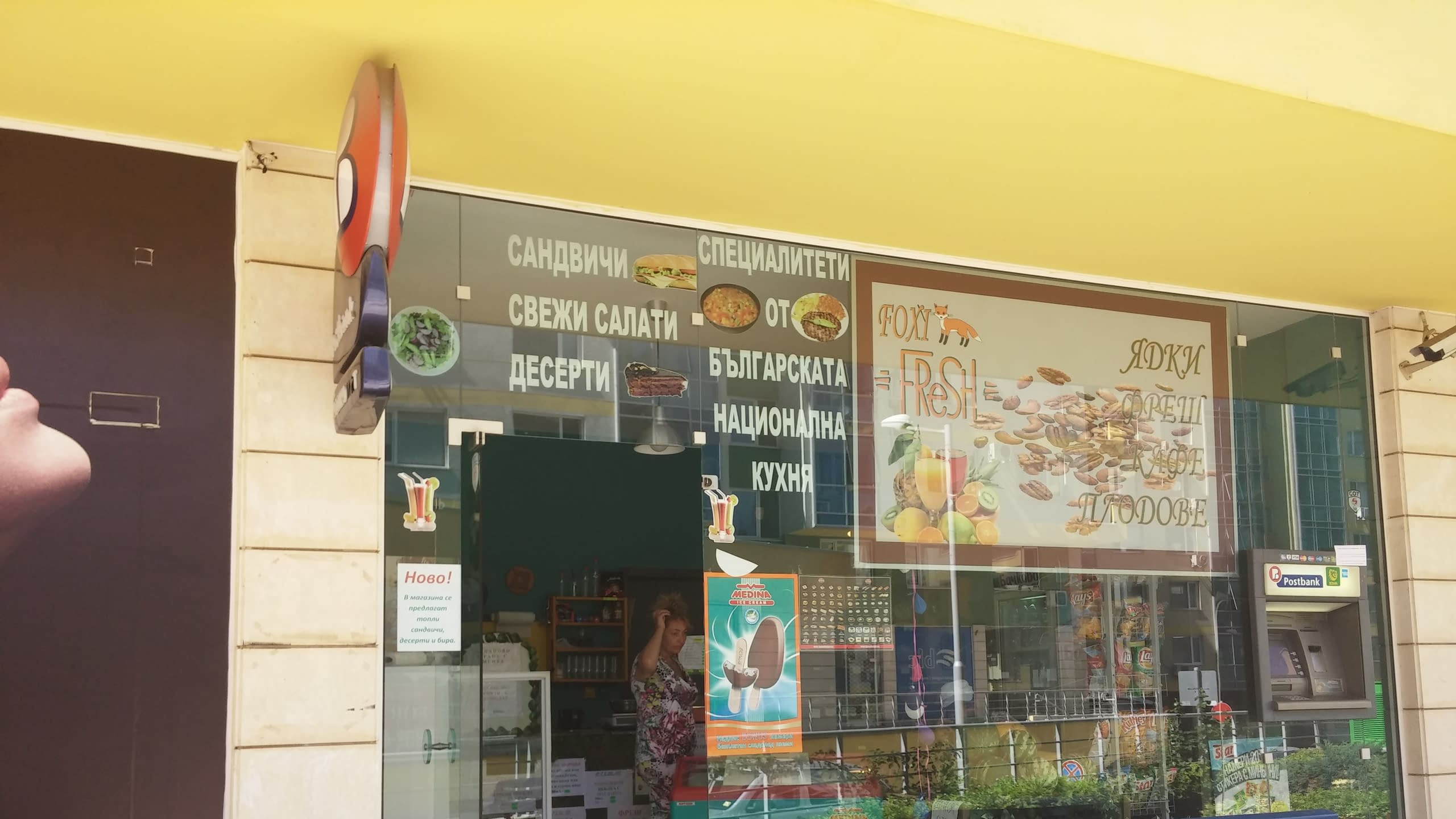 Вход на магазин за хранителни стоки, чиято стъклена витрина е облепена с различни стикери с надписи и изображения на храни.