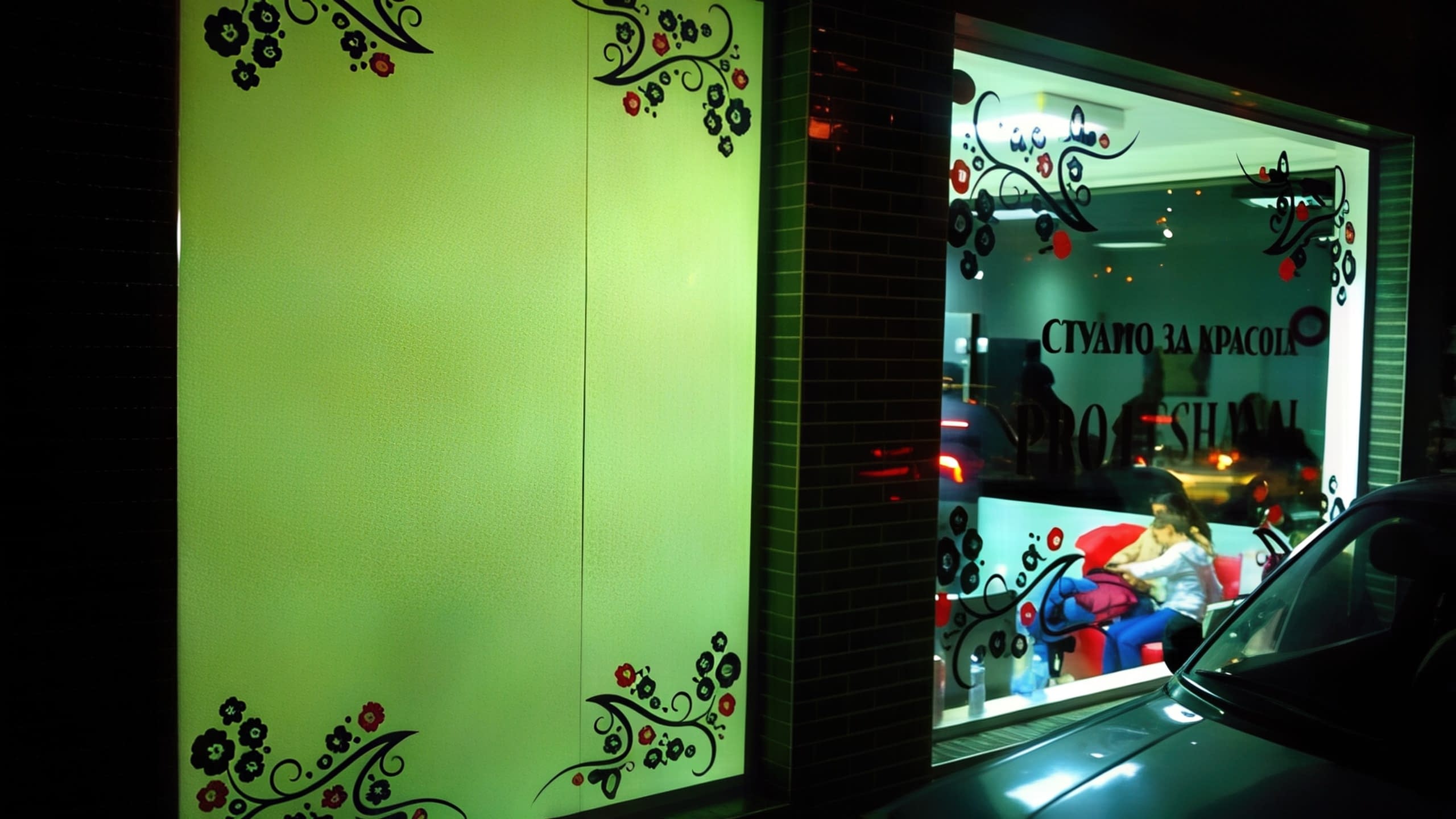 Външен изглед на салон през нощта, показващ една прозрачна и една матирана стъклена част, и двете с декоративни флорални стикери в ъглите.