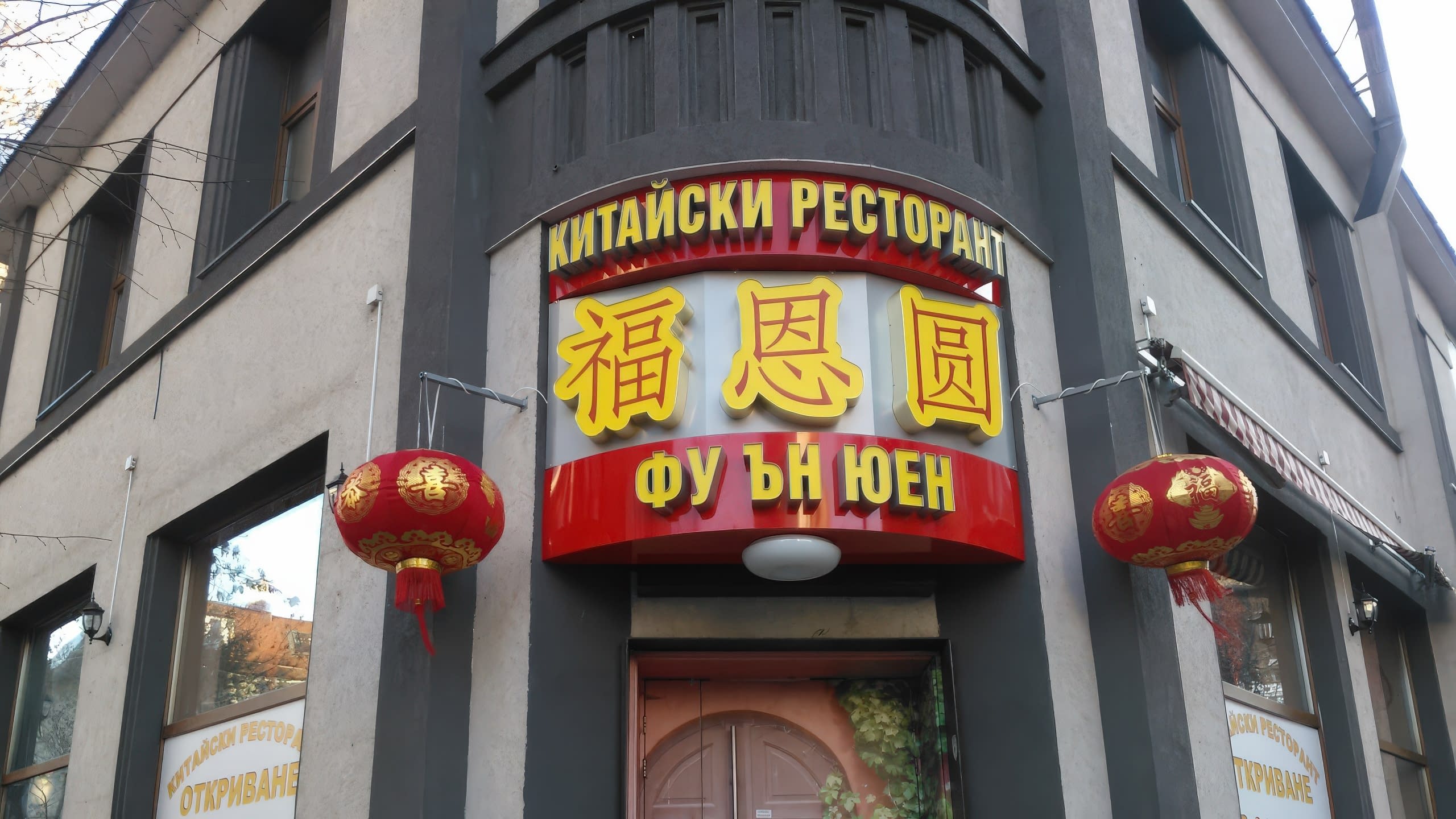 Извита табела, монтирана на ъгъла на сива сграда, с надписи на кирилица и китайски йероглифи, заобиколена от два червени фенера.