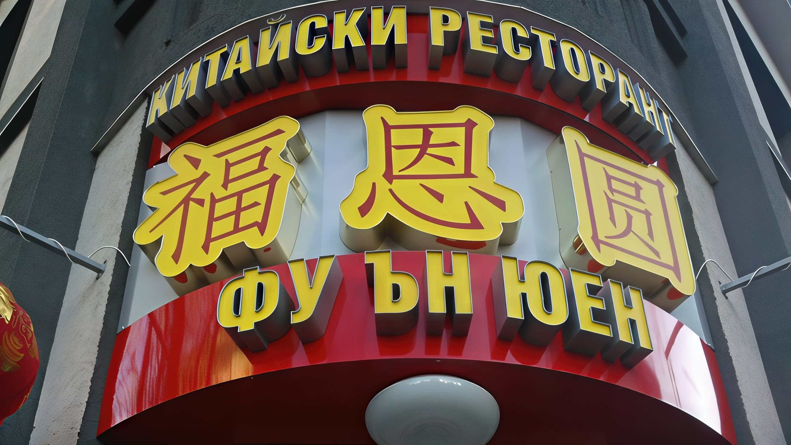 Изглед отблизо и под ъгъл към табела с триизмерни жълти букви и йероглифи, монтирани върху червена извита основа на фасада.