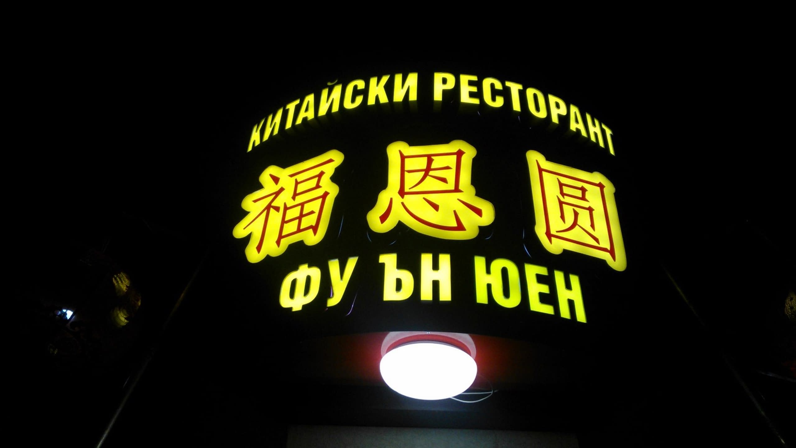 Осветена табела на ресторант през нощта, със светещи жълти букви на кирилица и китайски йероглифи на черен фон.