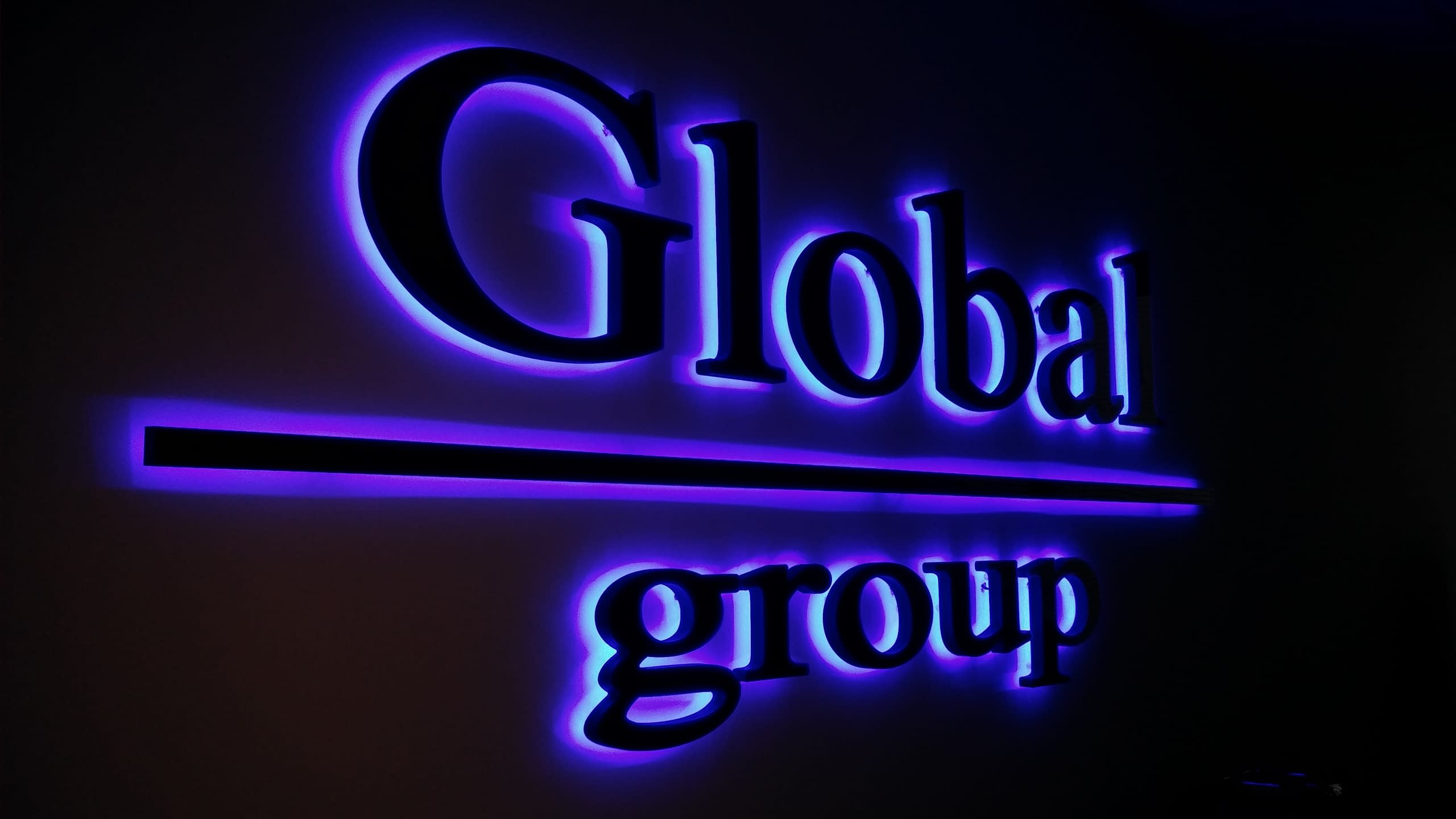 Светещ в лилаво надпис „Global group“, монтиран на стена в тъмно помещение.