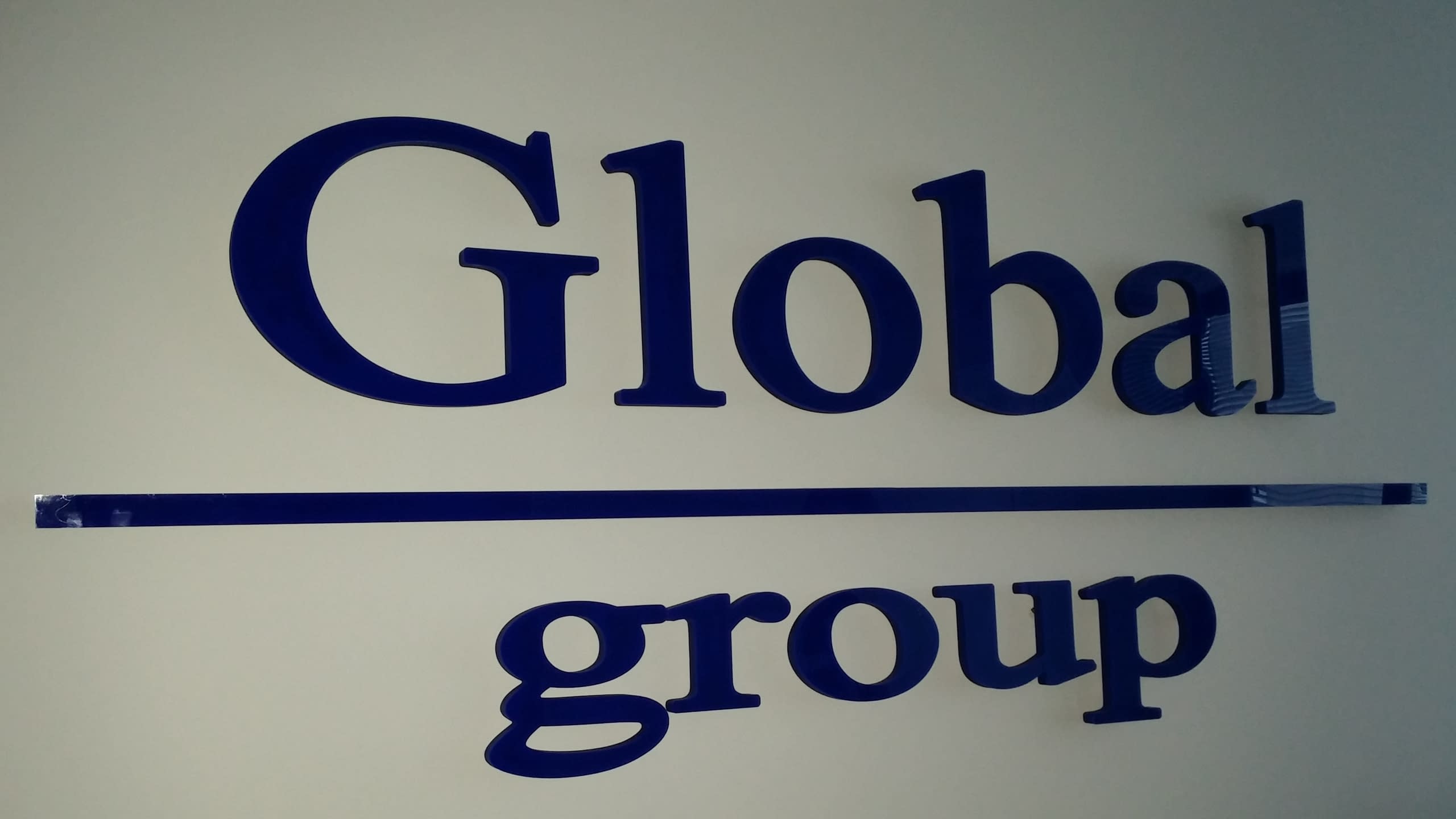Близък план на несветещ надпис „Global group“ от тъмносини обемни букви на светла стена.