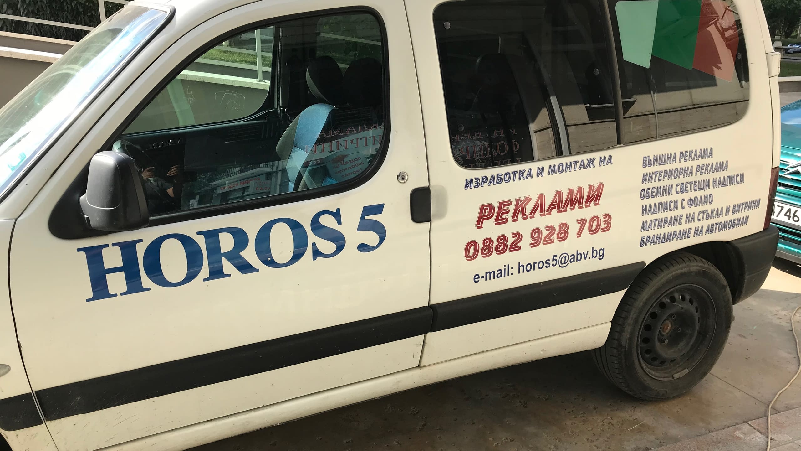 Страничен изглед на бял служебен автомобил с надписи в синьо и червено, които указват име на фирма, контакти и списък с дейности.