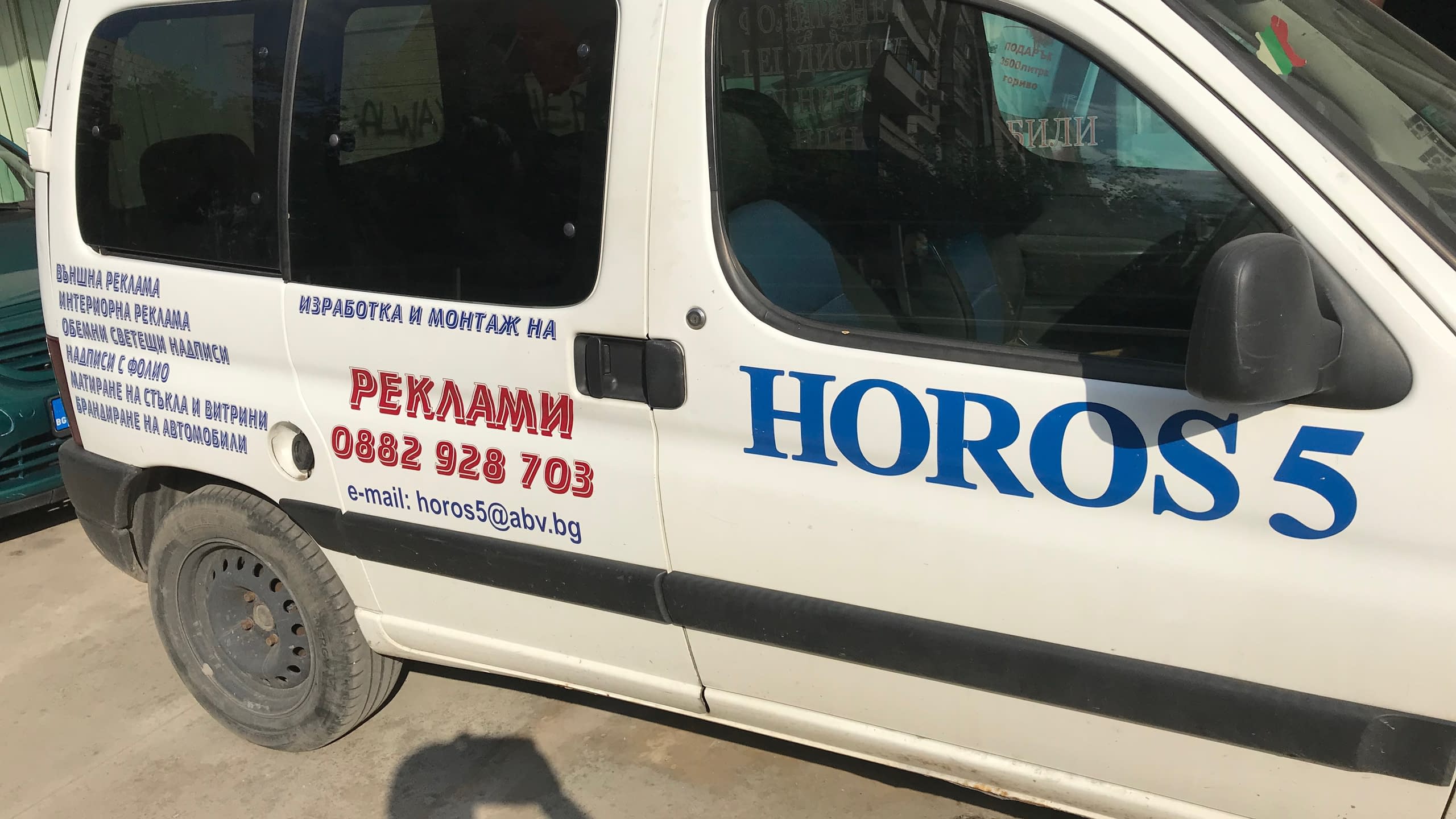 Бял автомобил, заснет отстрани, с апликирани стикери с името на фирма "HOROS 5" в синьо и друга информация за контакт в червено и черно.