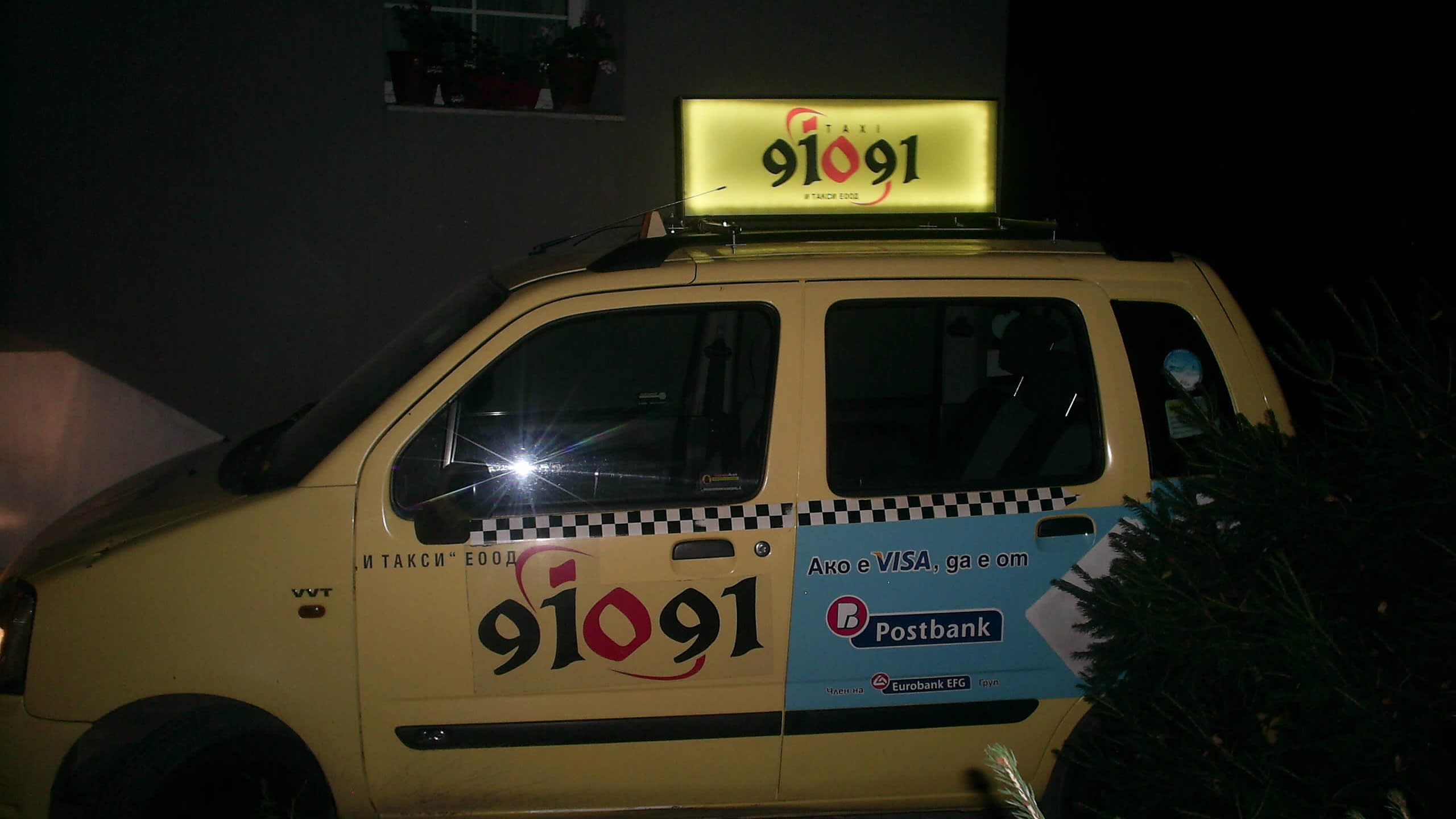 Брандиран таксиметров автомобил със светеща табела на покрива и рекламни стикери на вратите, заснет през нощта.