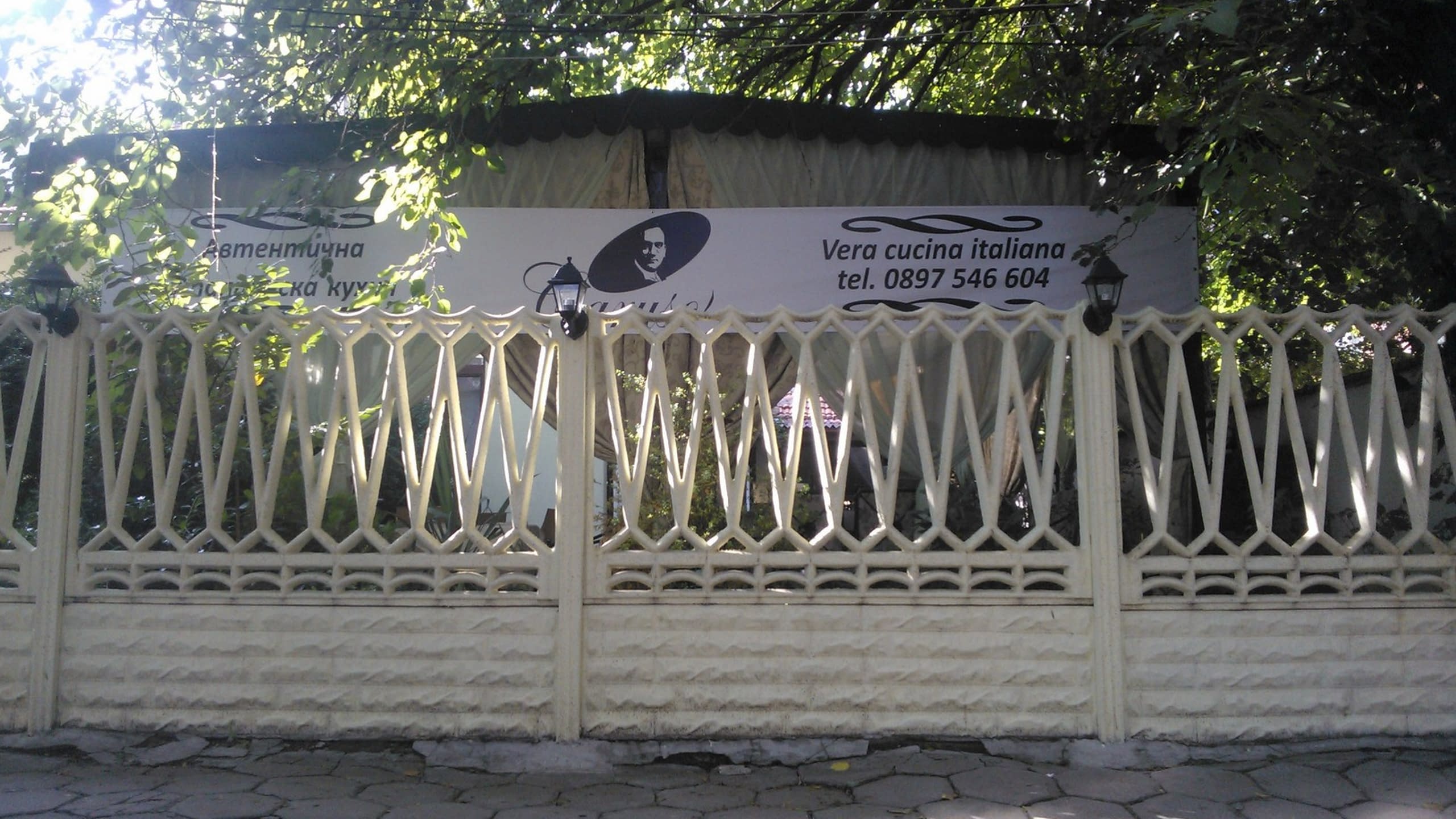 Голям бял транспарант за италиански ресторант, закачен на ограда пред градина със зеленина.