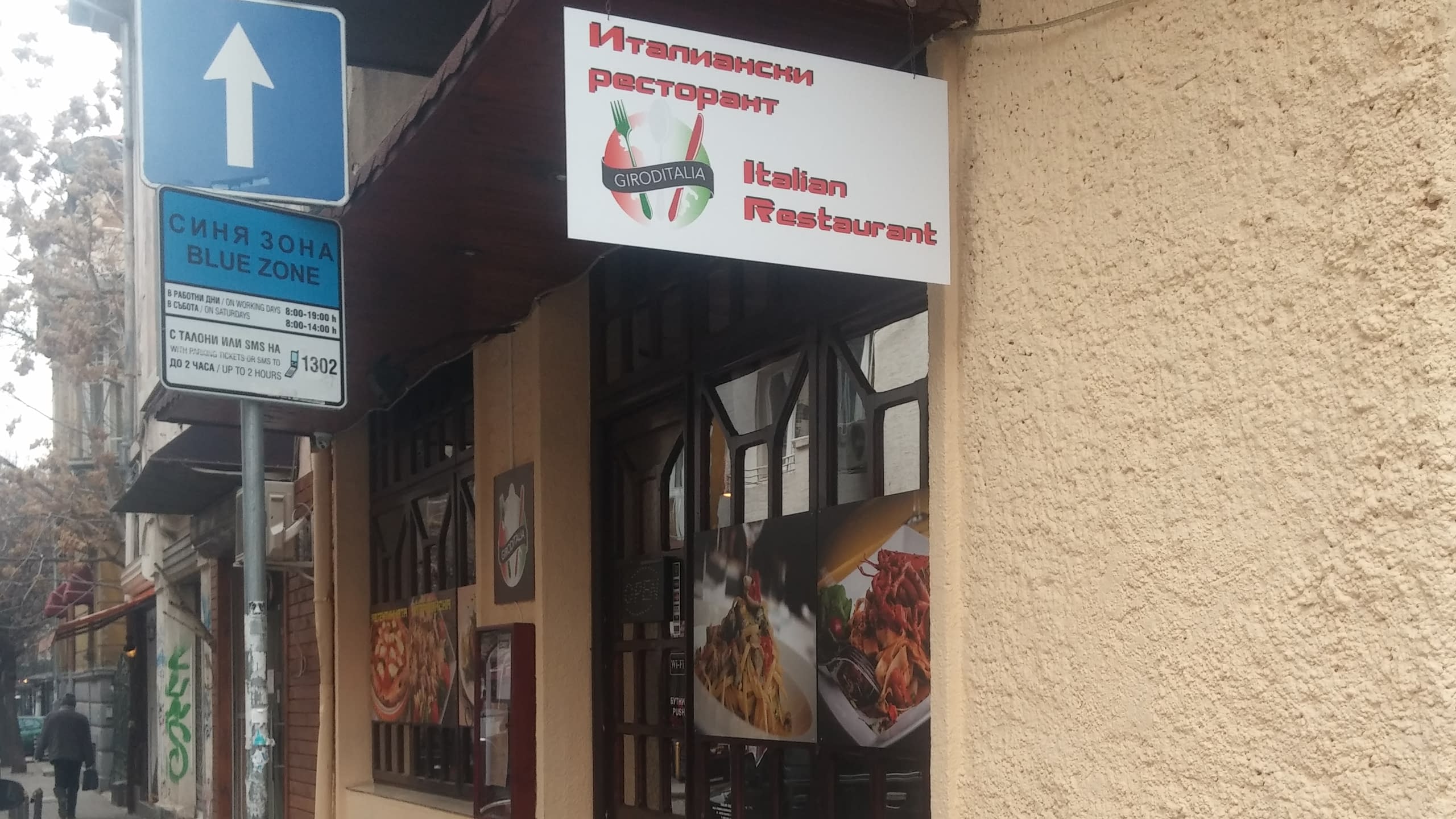 Страничен изглед на италиански ресторант с двуезична табела и пътен знак за "Синя зона".