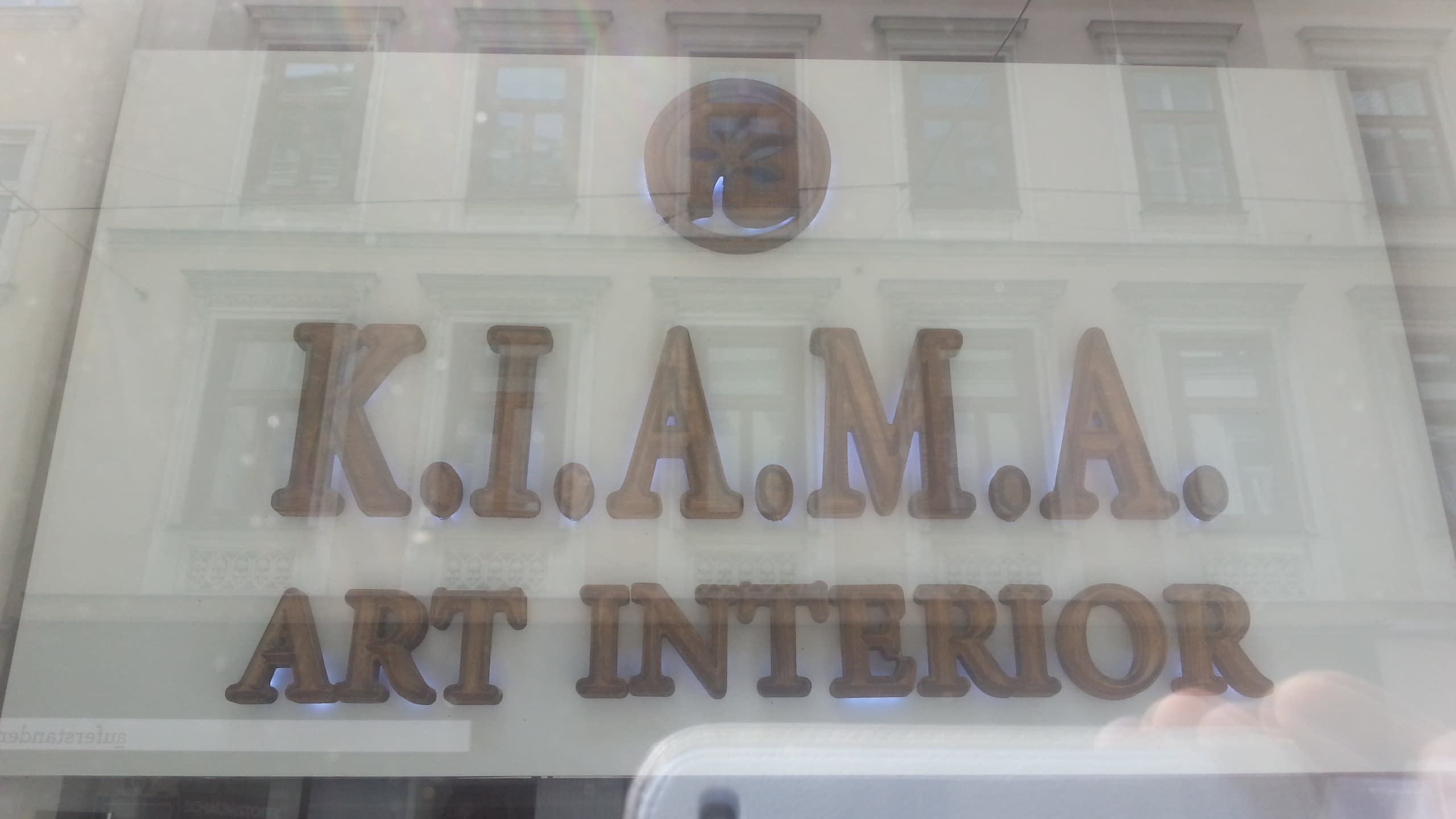 Снимка на надписа K.I.A.M.A., направенa през стъклото на витрина, в което се отразява сградата отсреща.