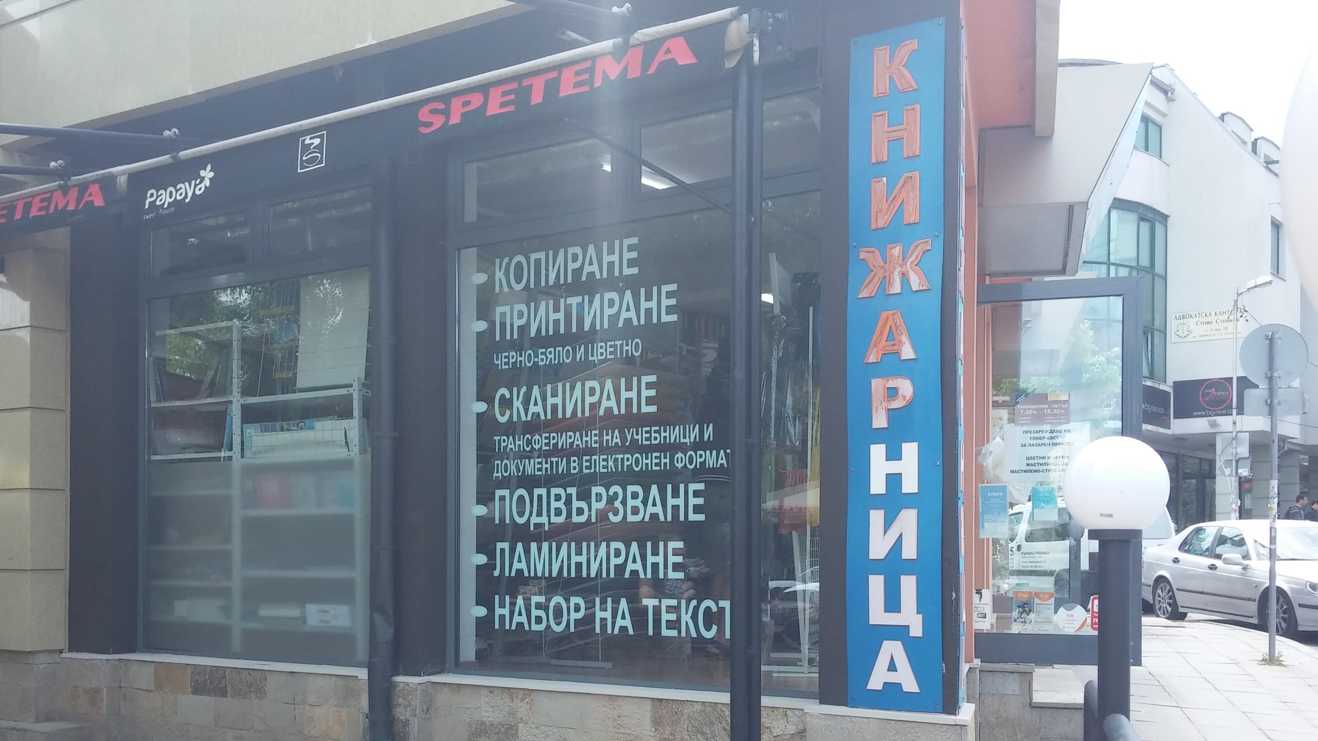 Външен изглед на книжарница "Спетема" с вертикална табела и списък с услуги от фолио на витрината.