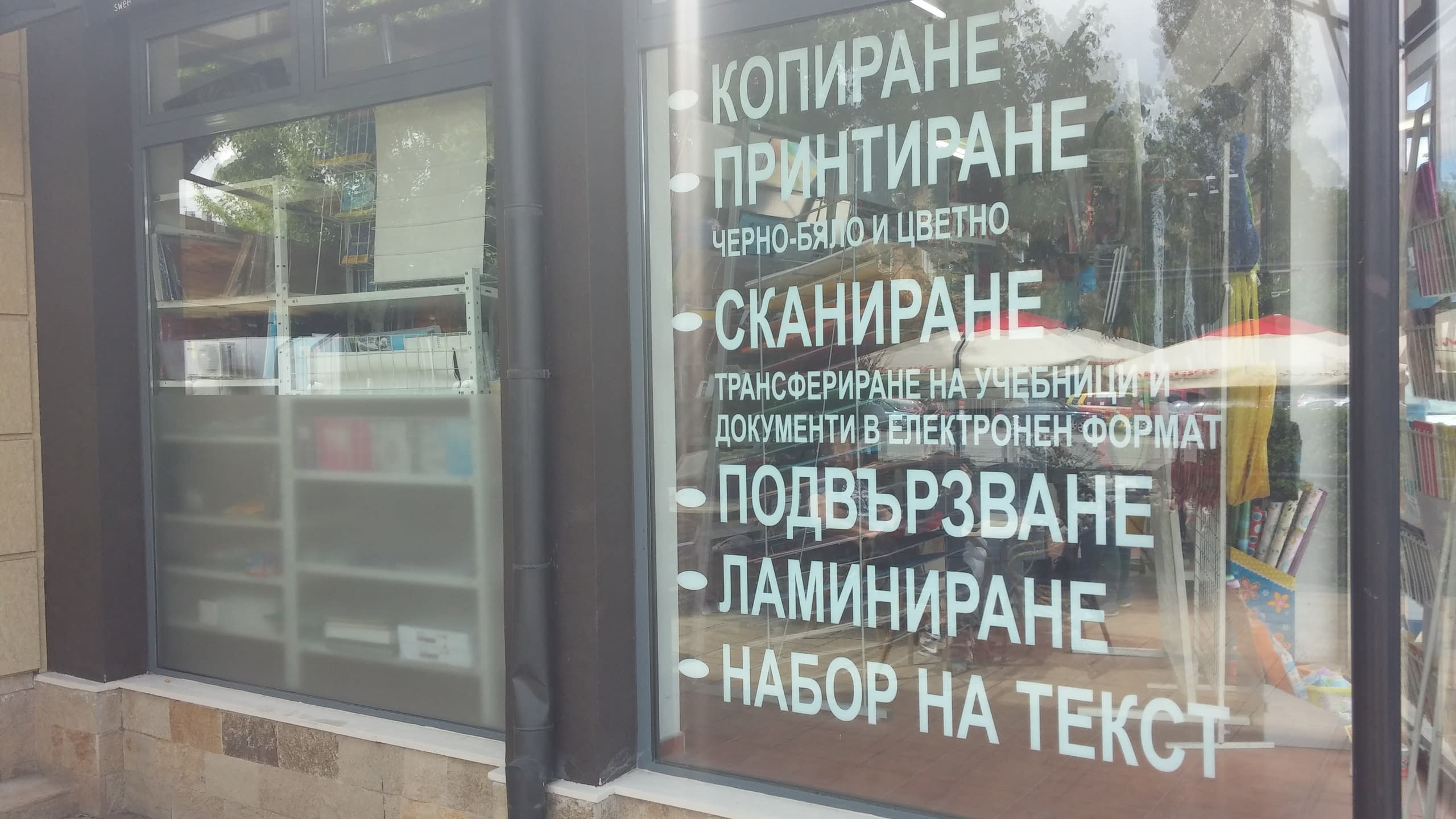 Списък с копирни услуги, изписан с бяло фолио върху стъклена витрина на книжарница.