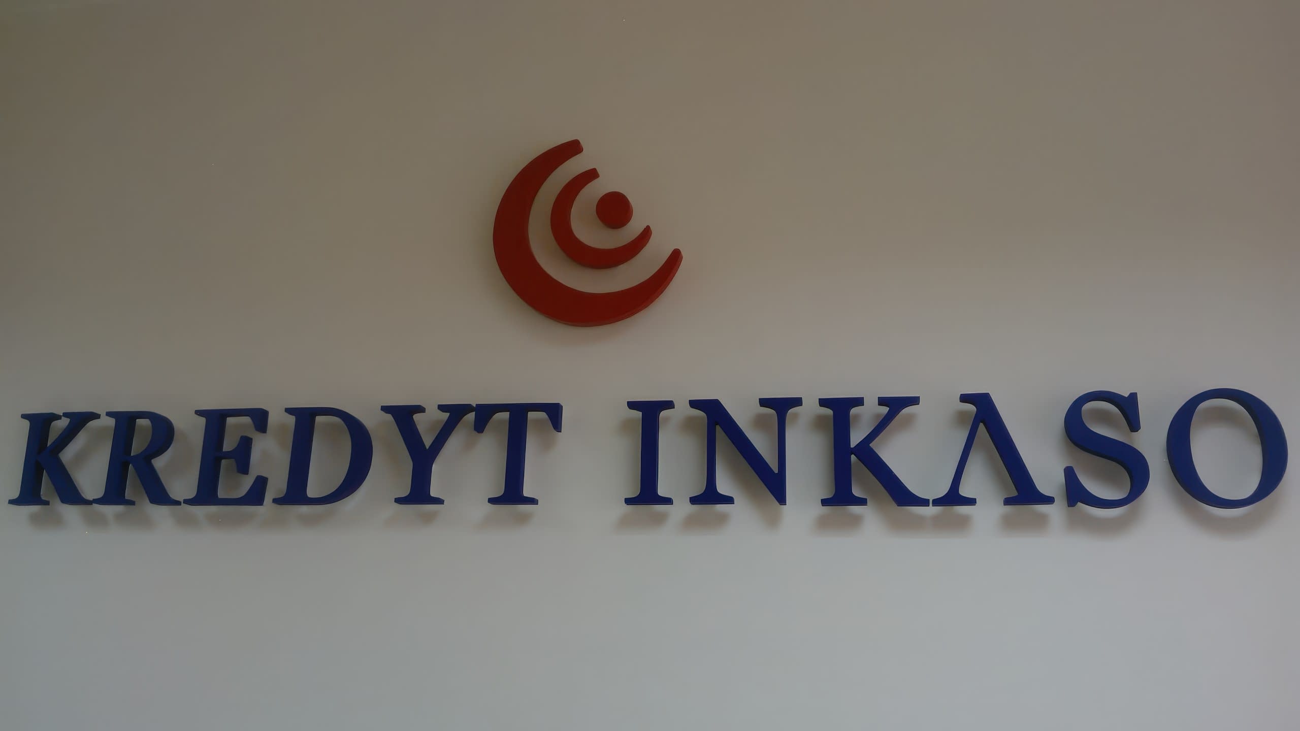 Фронтален изглед на сини букви с името KREDYT INKASO и червено лого, разположени централно на стена.