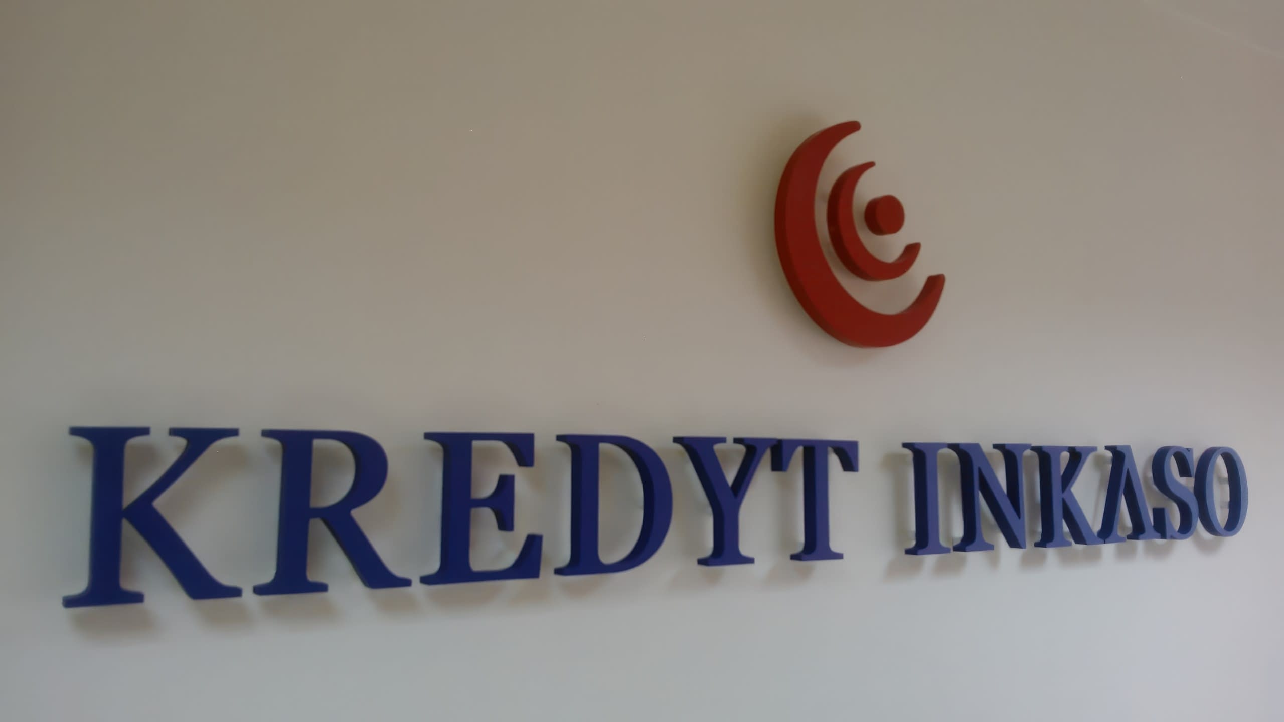 Червено лого и сини релефни букви на бяла стена в офис, заснети в близък план и леко отстрани.