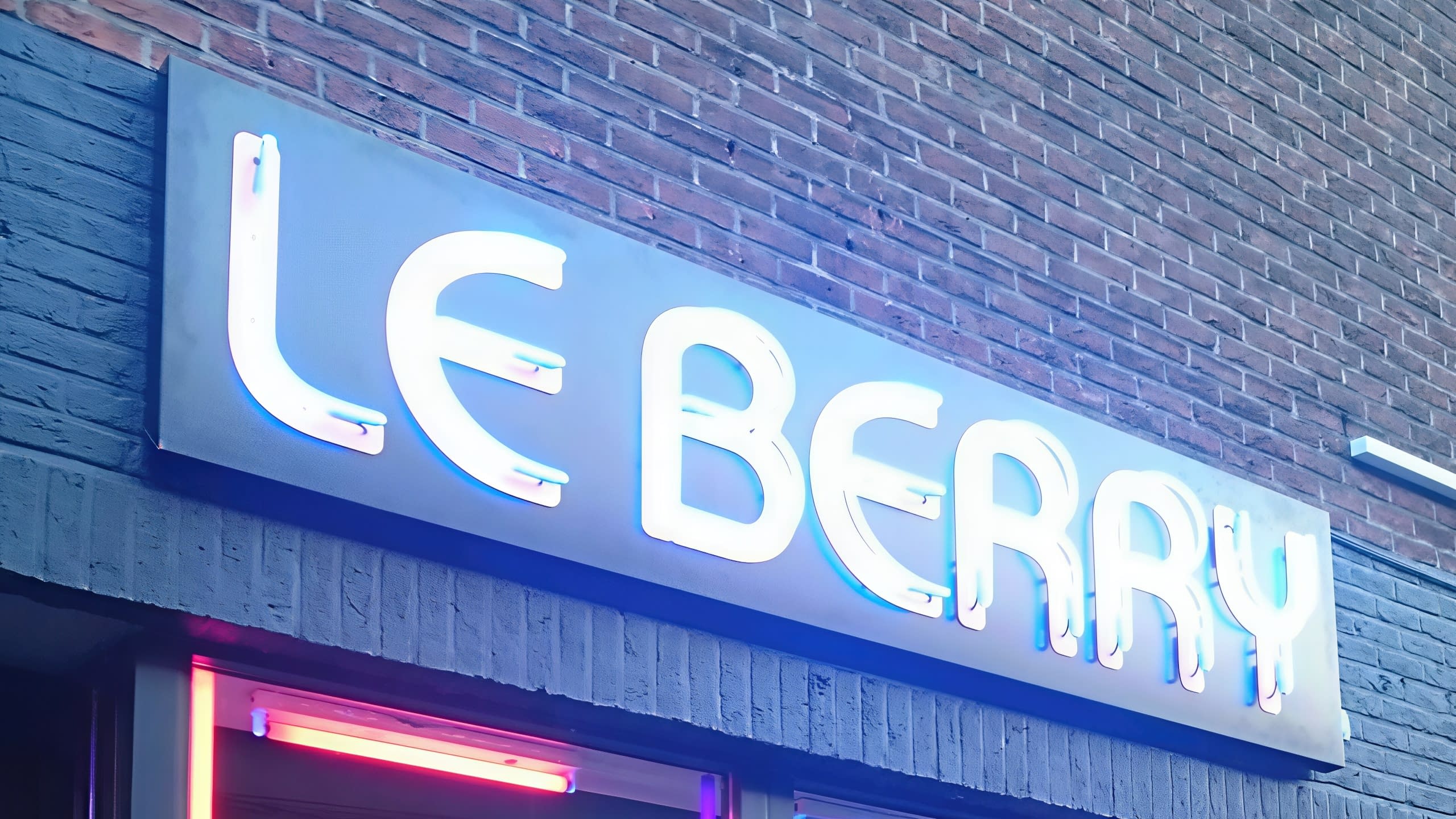 Надпис LE BERRY, светещ в бяло-синя светлина, монтиран на тъмна тухлена фасада и заснет отдолу.