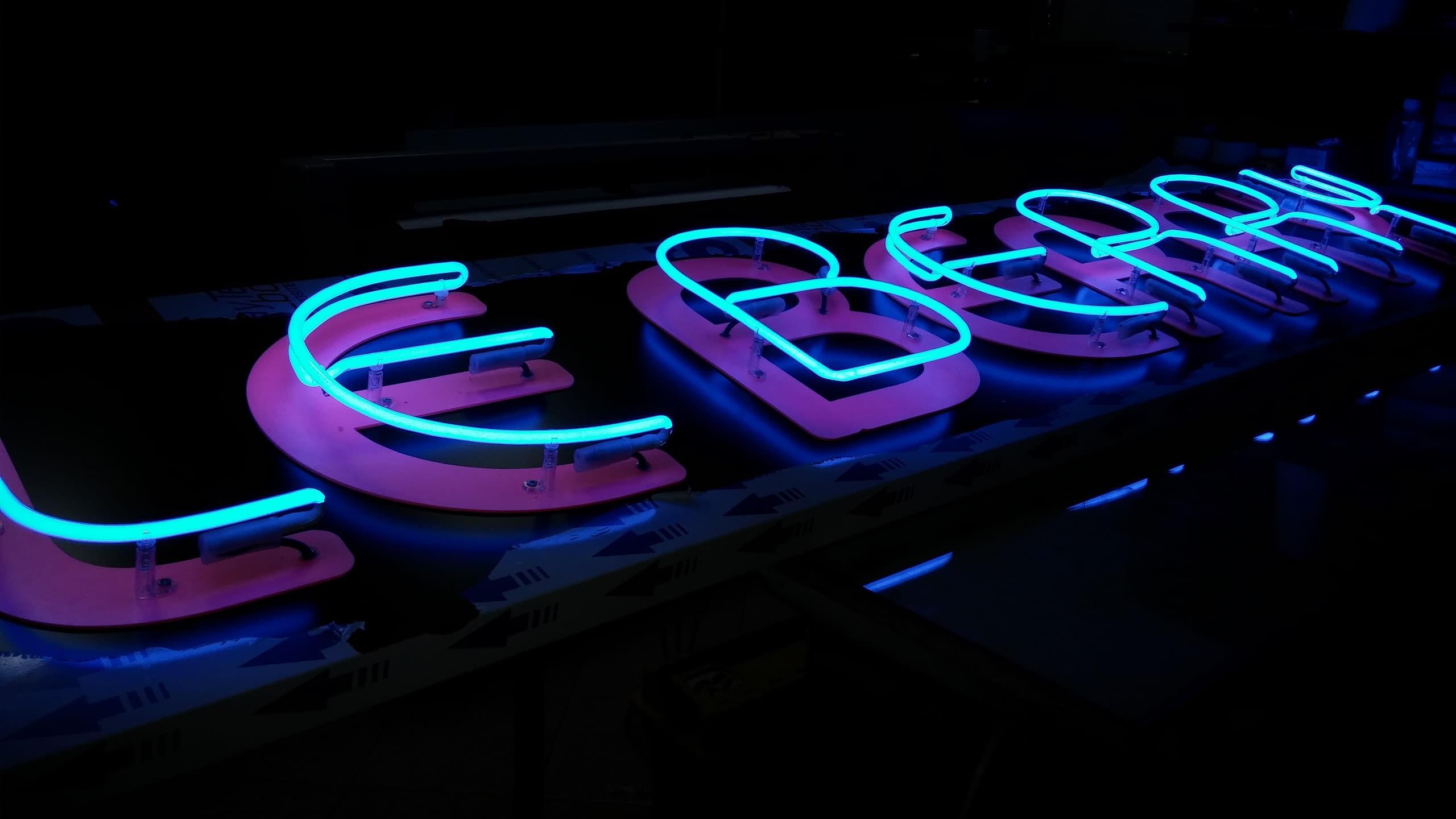 Светещ надпис със сини контури върху розови букви, заснет под ъгъл в тъмното преди финалния монтаж.