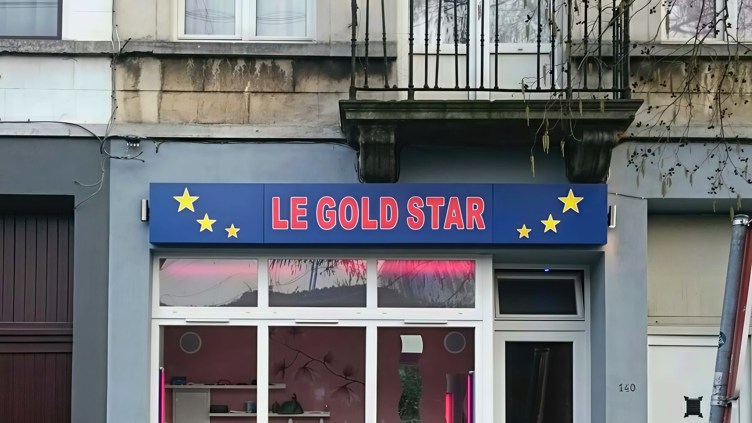 Синя правоъгълна табела с червен надпис LE GOLD STAR и жълти звезди, монтирана над витрина на салон през деня.