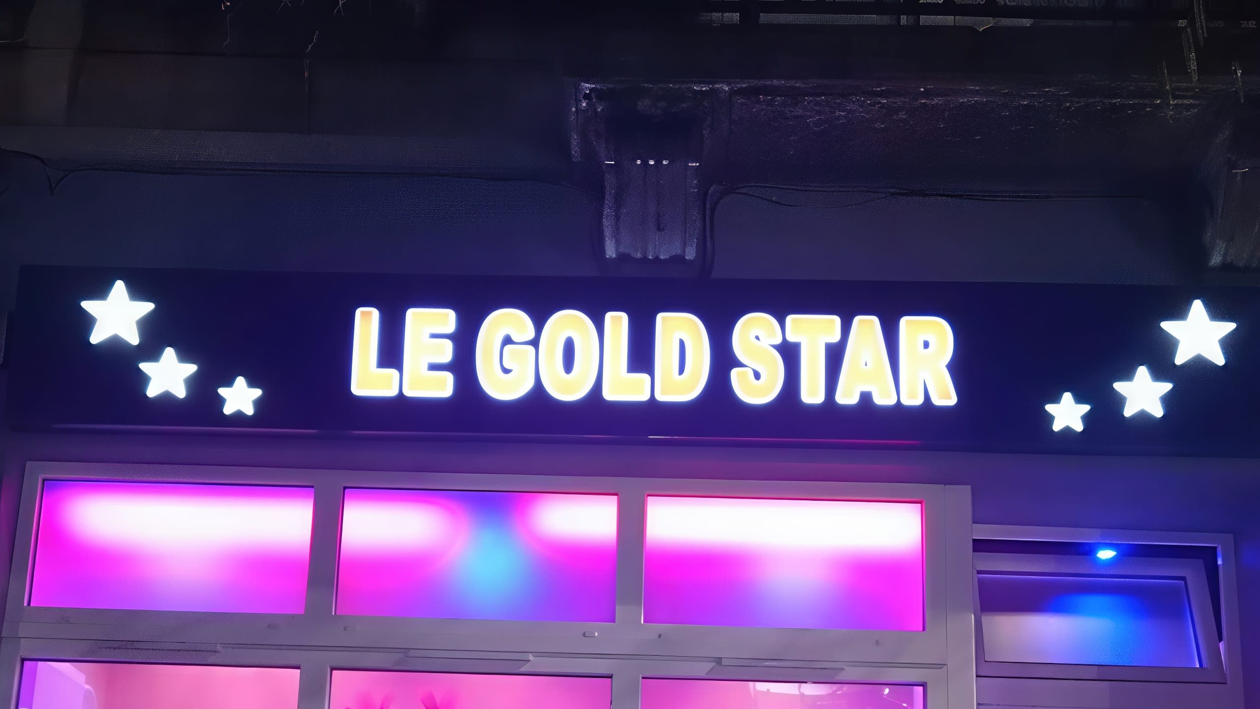 Светещи в бяло-жълто букви и звезди на надписа LE GOLD STAR, монтиран над витрина с цветни светлини през нощта.