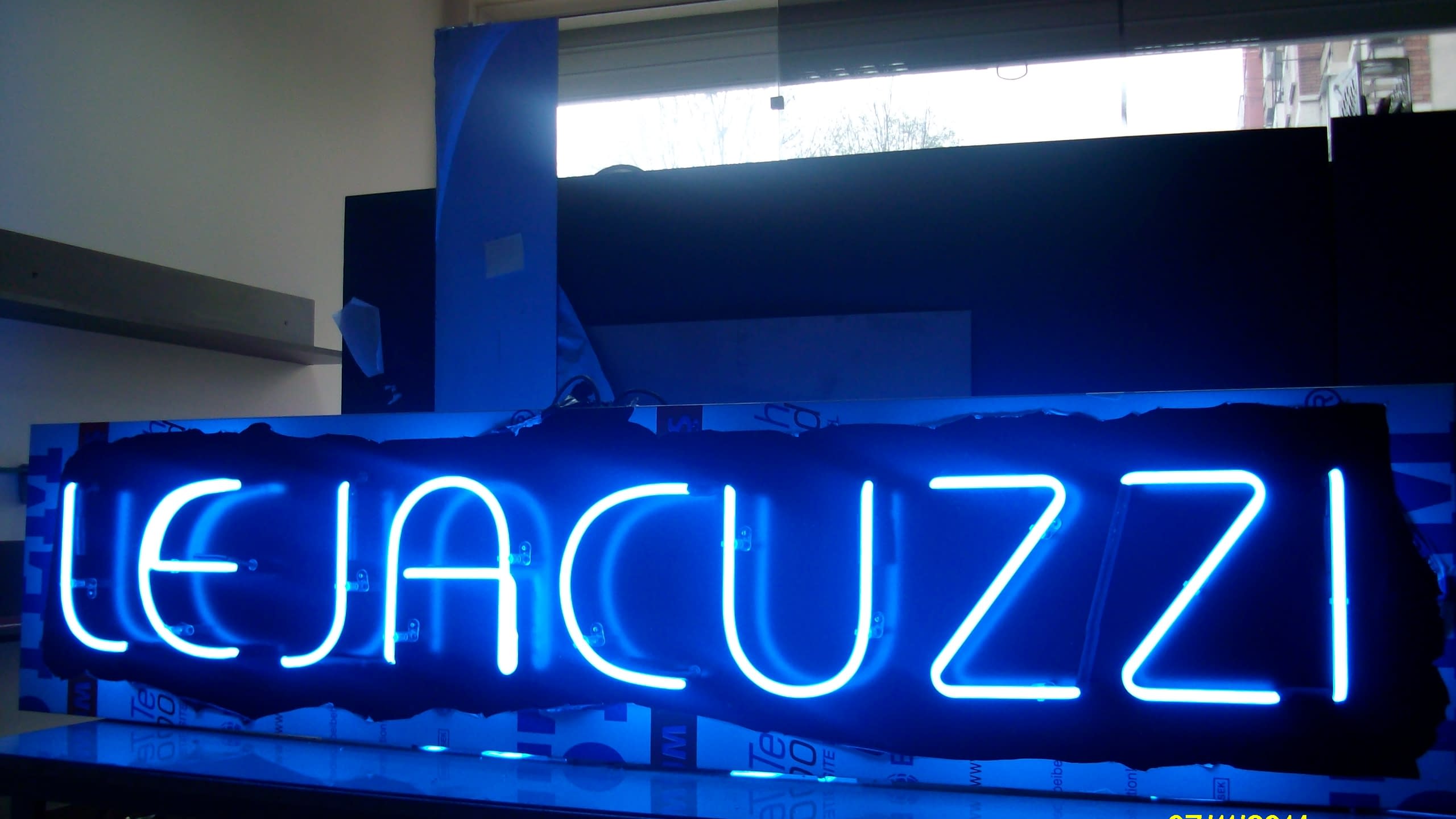 Надпис LE JACUZZI, светещ в наситено синьо, заснет в близък план с отражение на светлината върху повърхността под него.