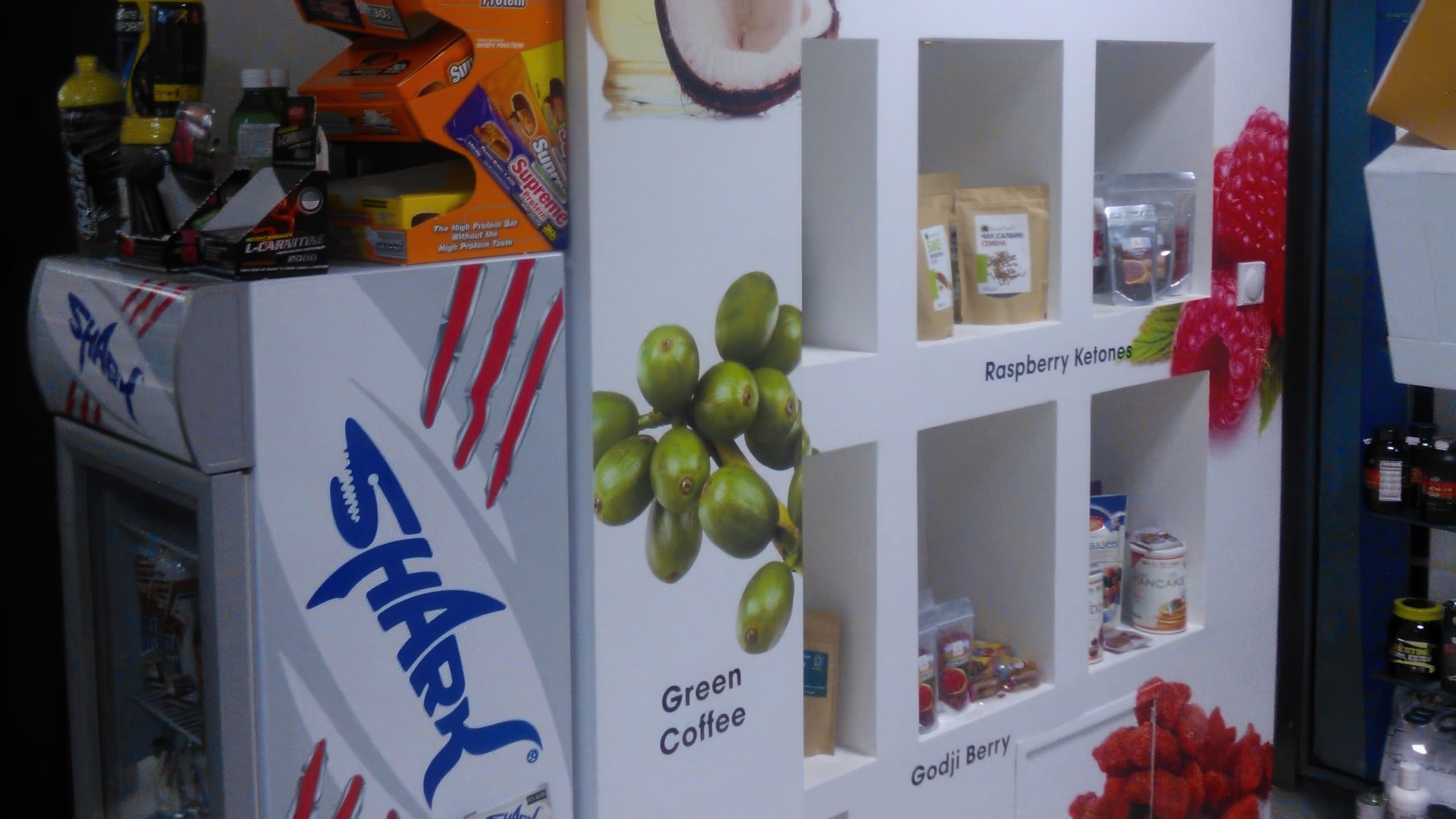 Близък план на рафтове със здравословни храни, декорирани със стикери на зелено кафе и годжи бери, до хладилник с напитки.