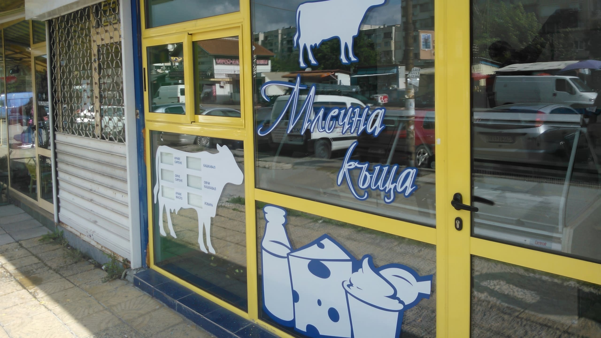 Витрина на млечен магазин с жълти дограми и стикери от фолио с форма на крава и сирене.