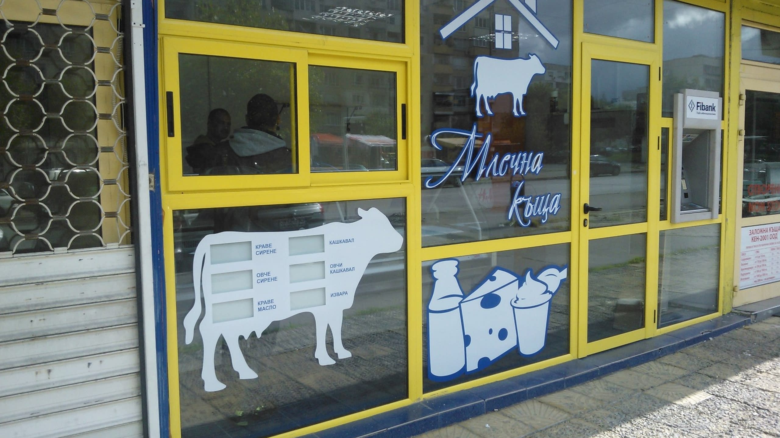 Цялостен изглед на витрина на магазин 'Млечна къща', брандирана със стикери на крави и млечни продукти.