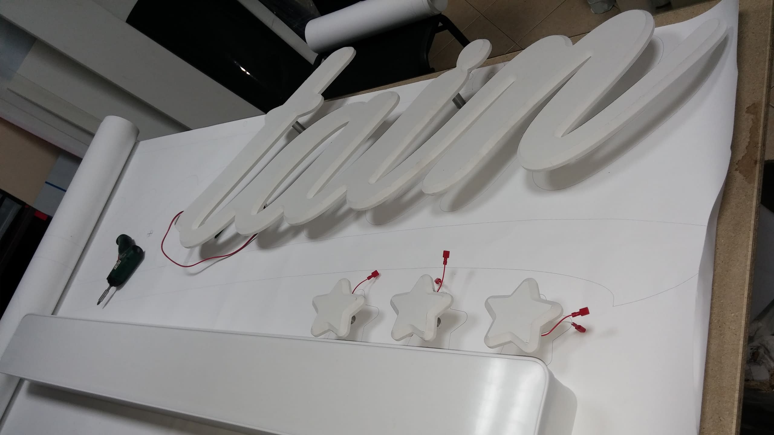 Бели обемни букви и звезди, положени върху хартиен шаблон в работилница, с видими кабели за електрическо свързване.