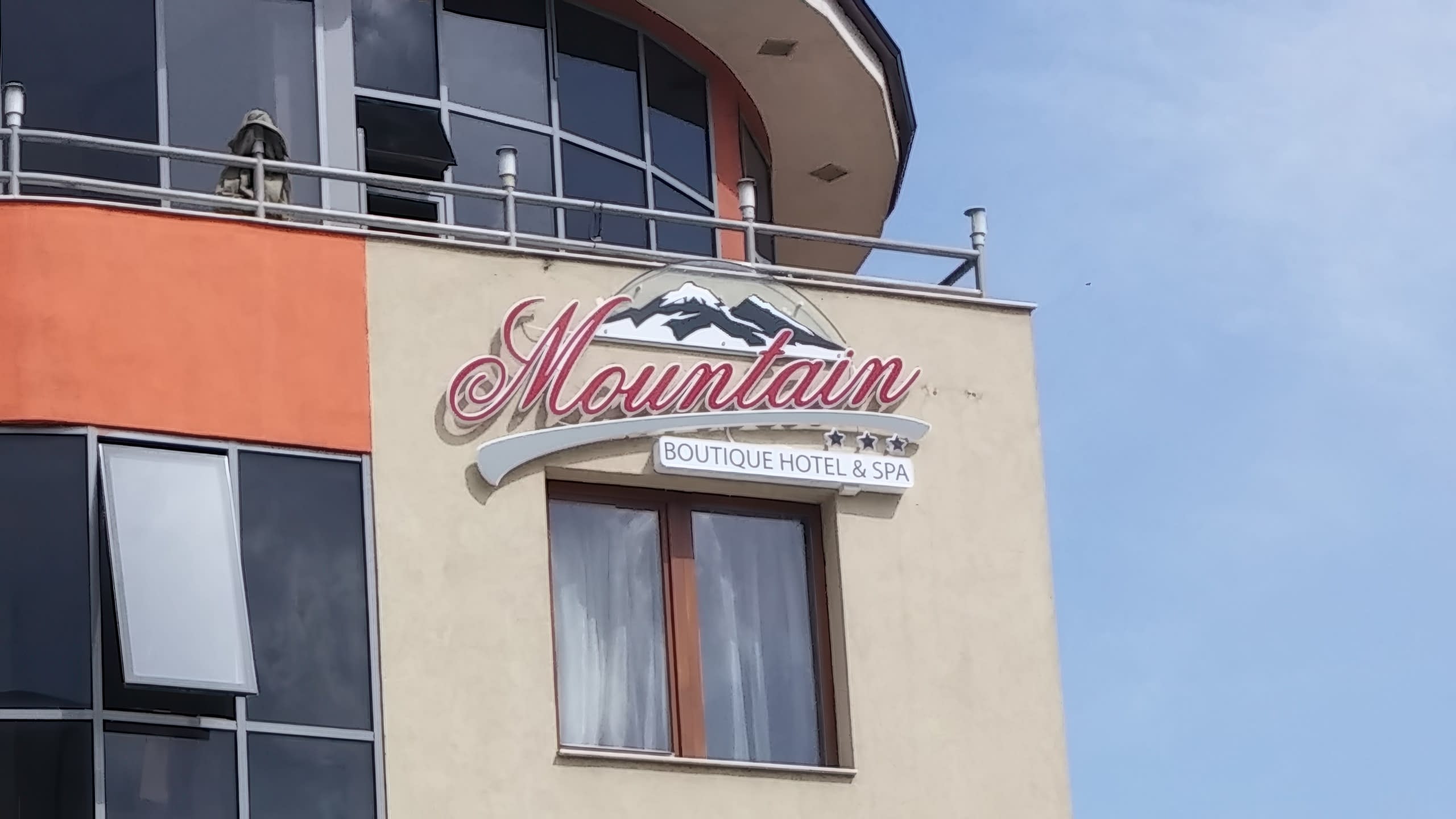 Табела на хотел, монтирана на бежова фасада под балкон със стъклен парапет.