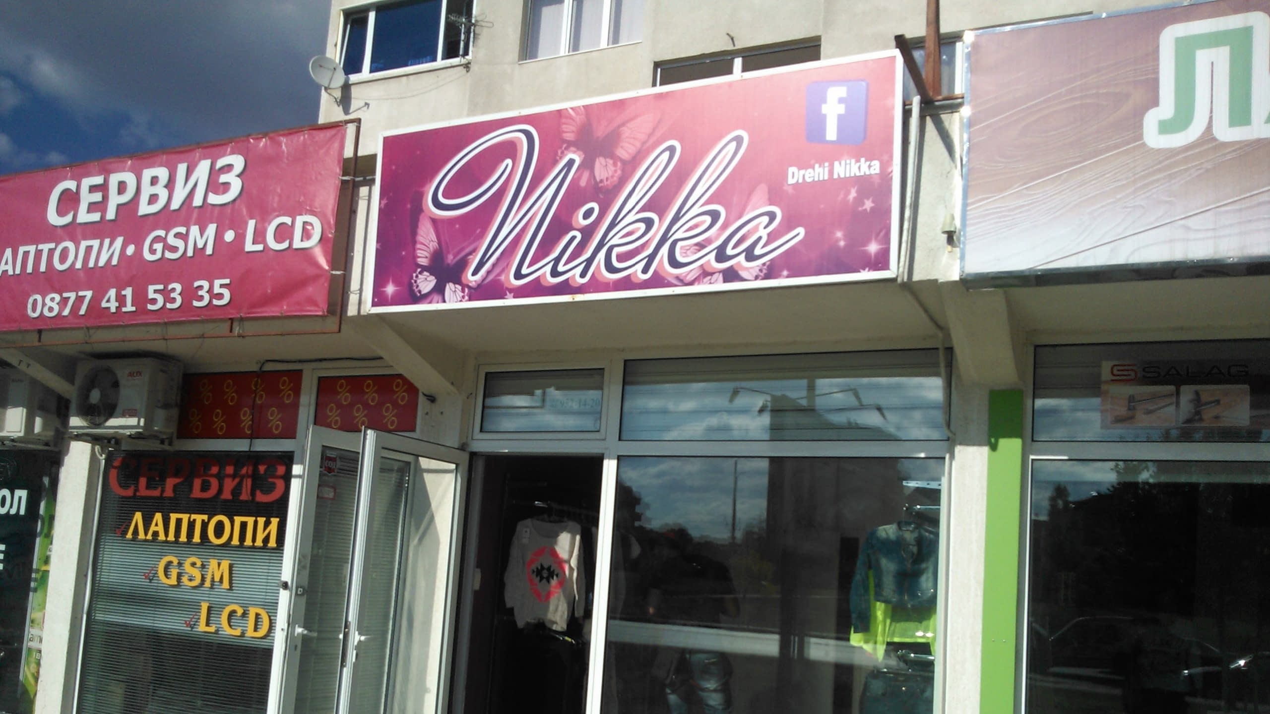 Табела с надпис "Nikka" над магазин за дрехи, заснета в слънчев ден, до съседен магазин за сервиз на техника.