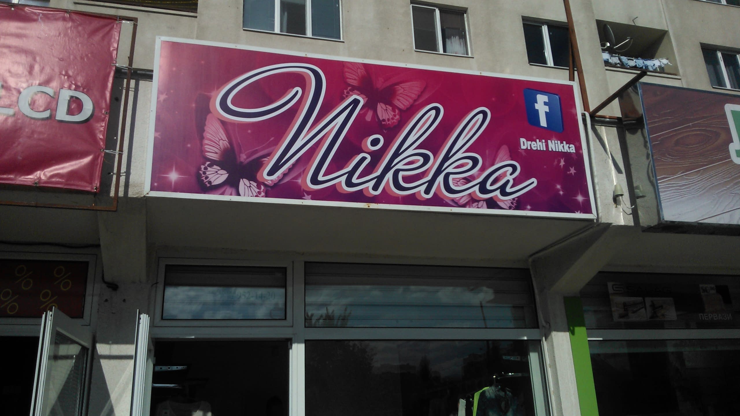 Розов банер с калиграфски надпис "Nikka" и пеперуди, монтиран над витрина на магазин, разположен на партерен етаж на жилищна сграда.