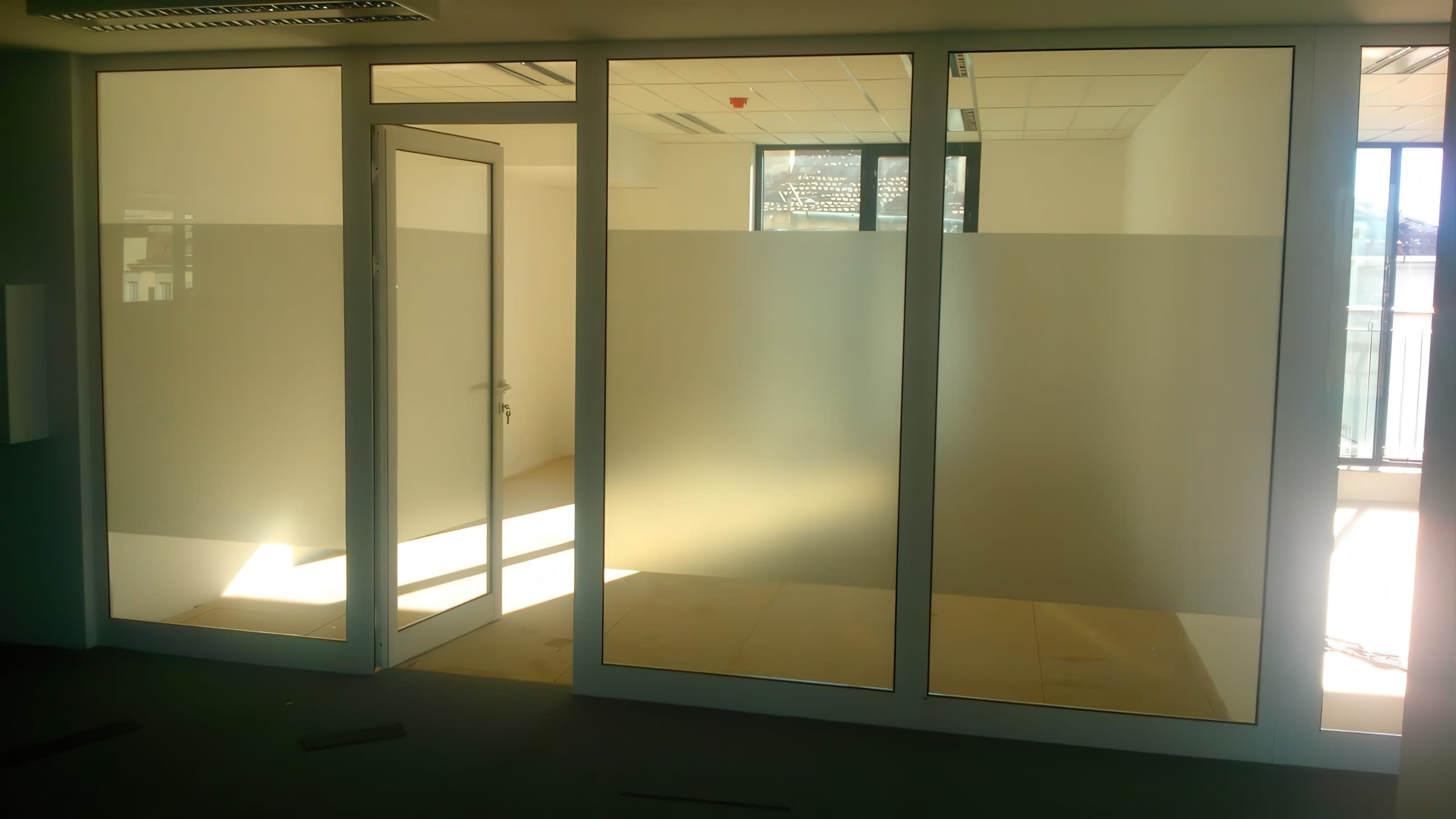 Офис преграда от стъкло и алуминий с матирано фолио по средата, гледана от тъмен коридор.