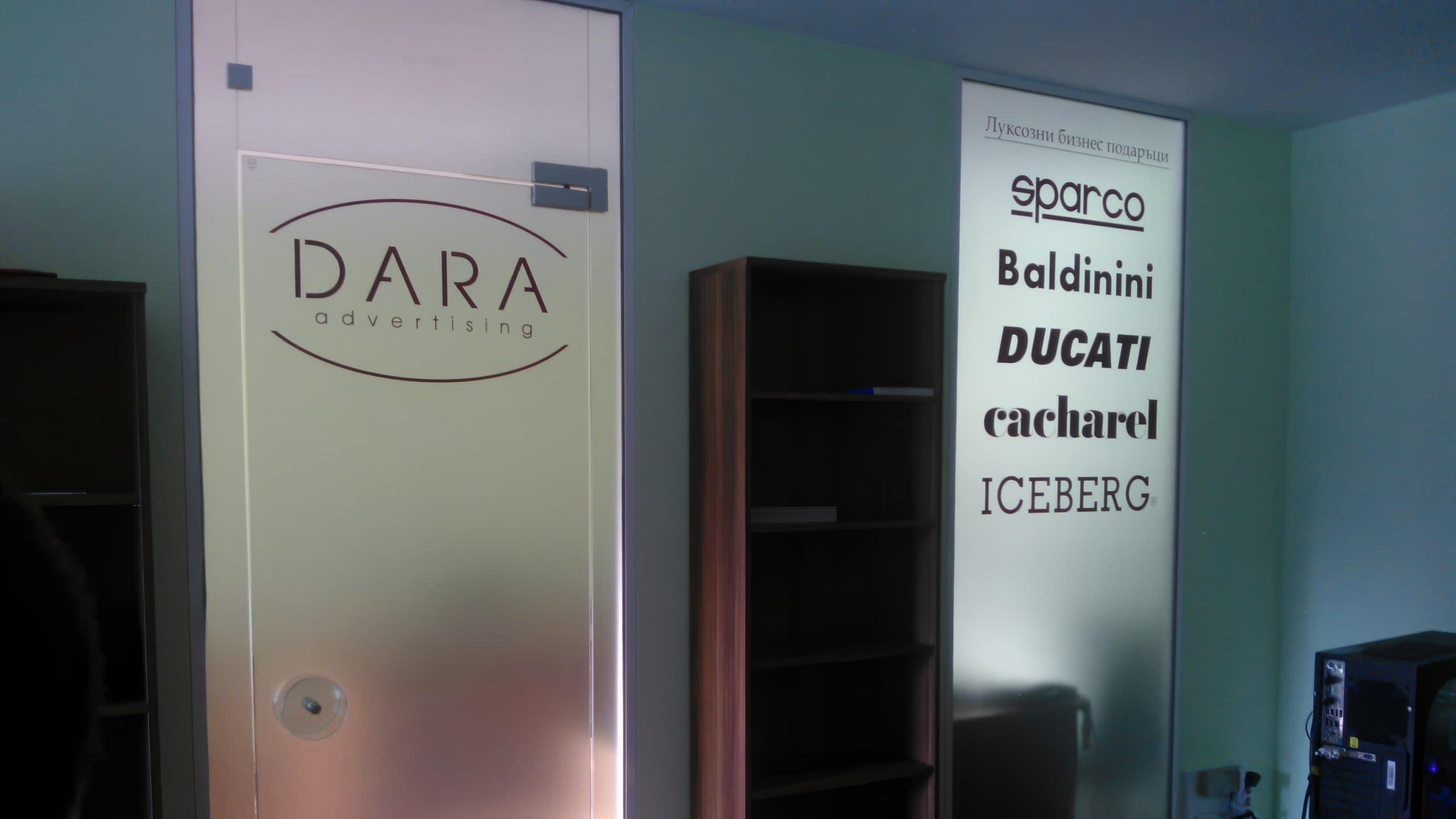 Матирана стъклена врата с лого "DARA advertising" до светещ панел с лога на луксозни марки.