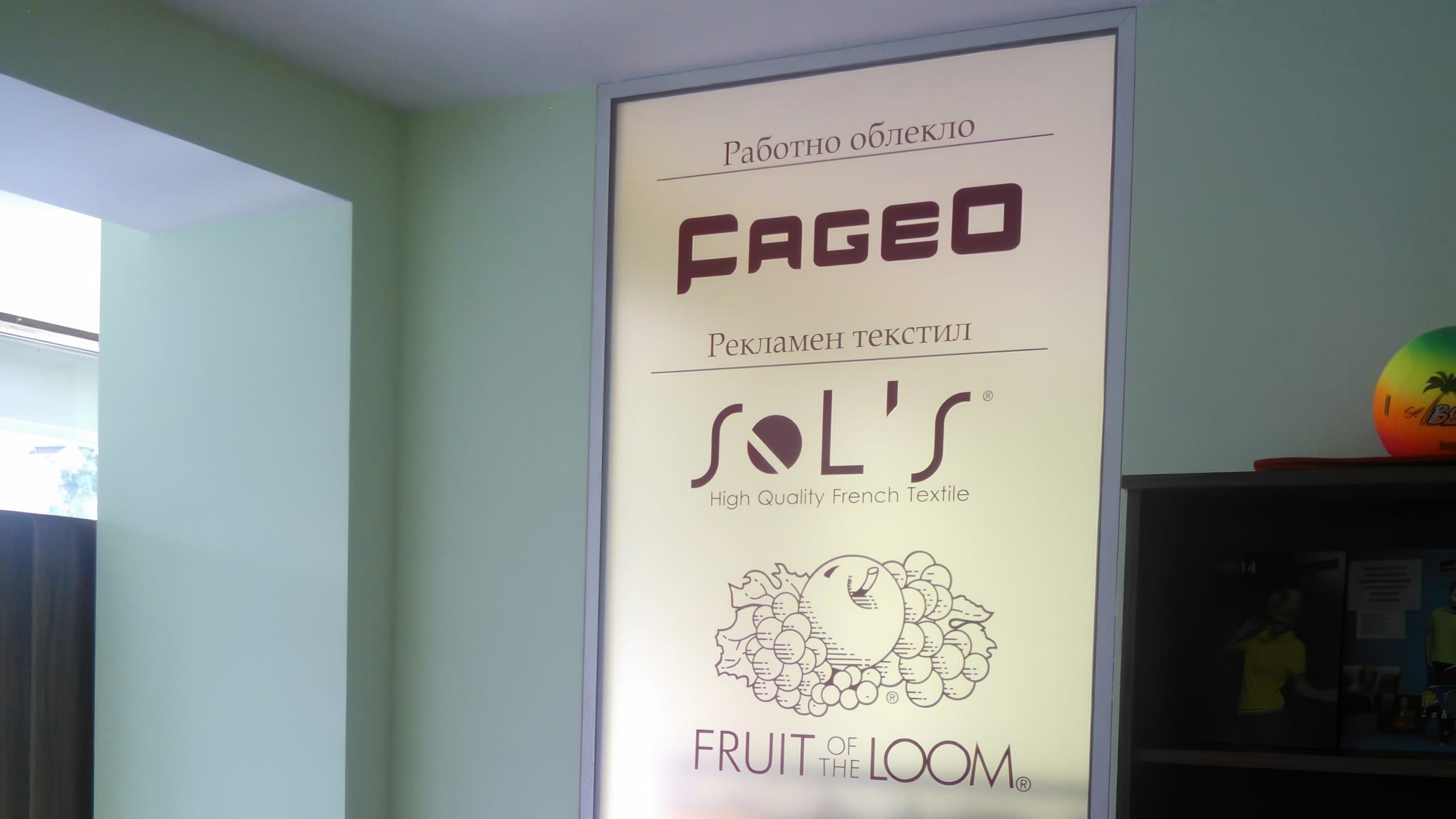 Светещ рекламен панел в офис, показващ лога на фирми за текстил като FAGEO и Fruit of the Loom.