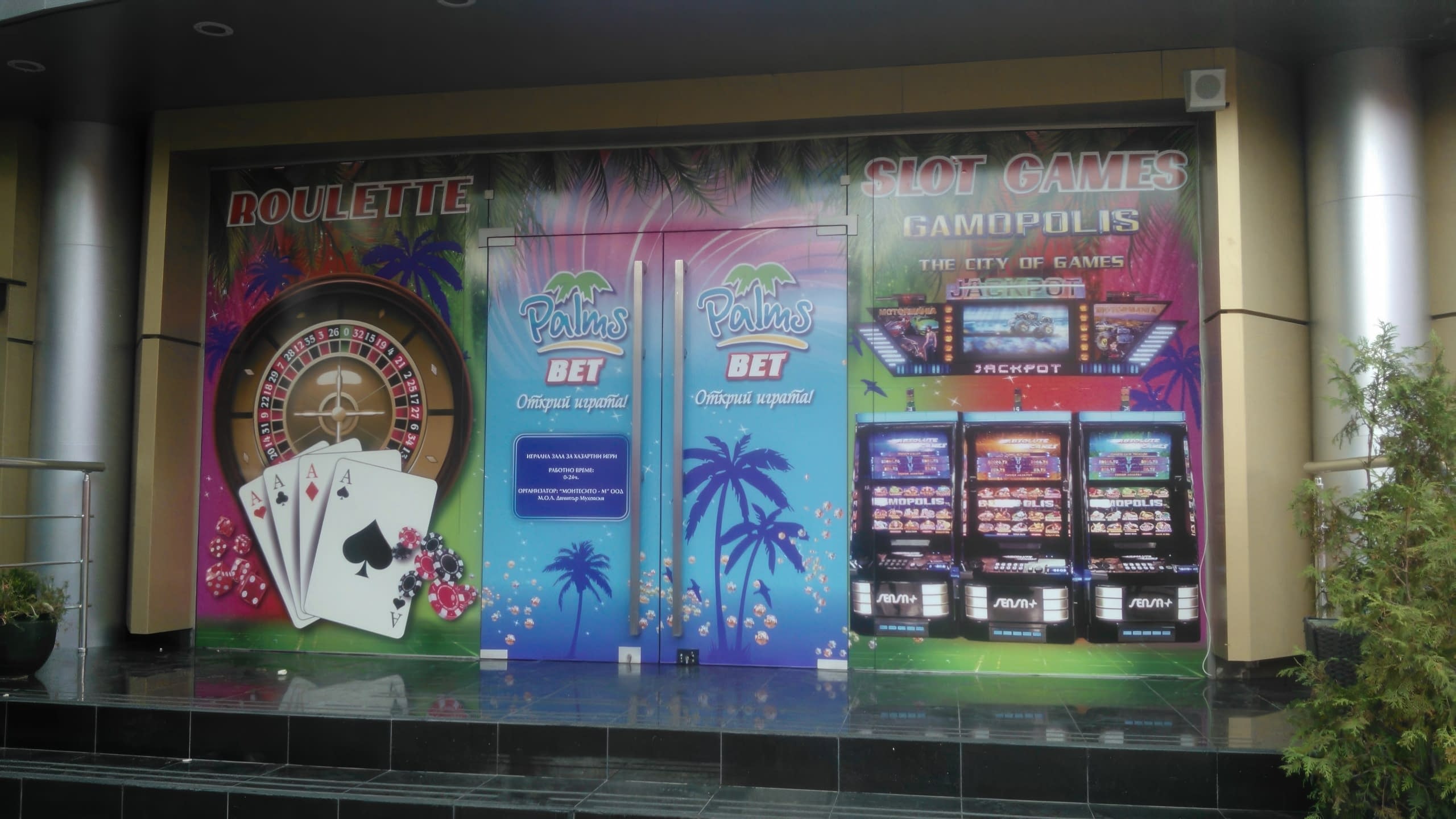 Изглед към входа на игрална зала Palms Bet, декориран с фолио с палми, рулетка и слот машини, заобиколен от декоративни растения.