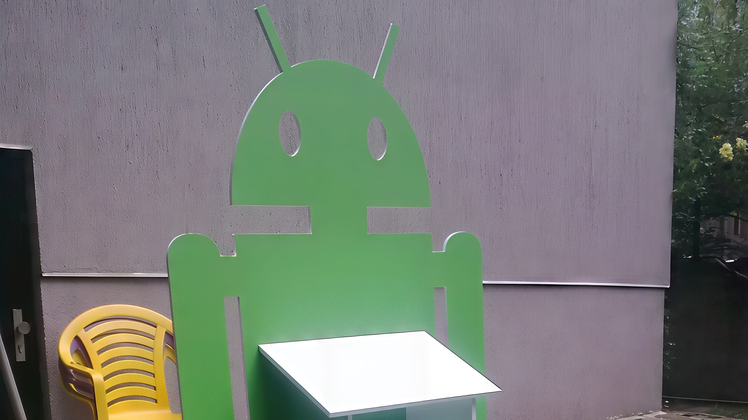 Голям зелен дисплей с формата на робот и бял рафт за продукти, поставен на открито до сива стена.