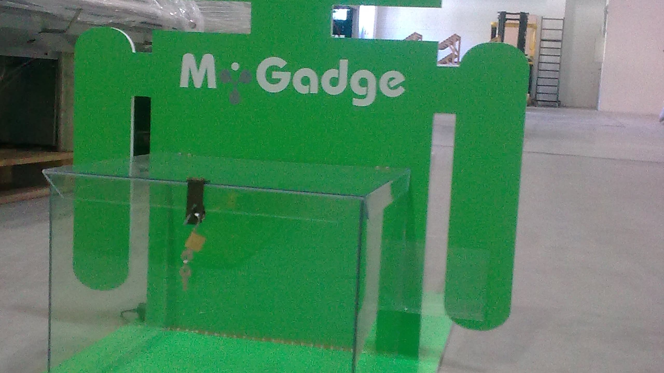 Горната част на зелен дисплей с форма на робот и надпис "M-Gadge", заснет в складово помещение.