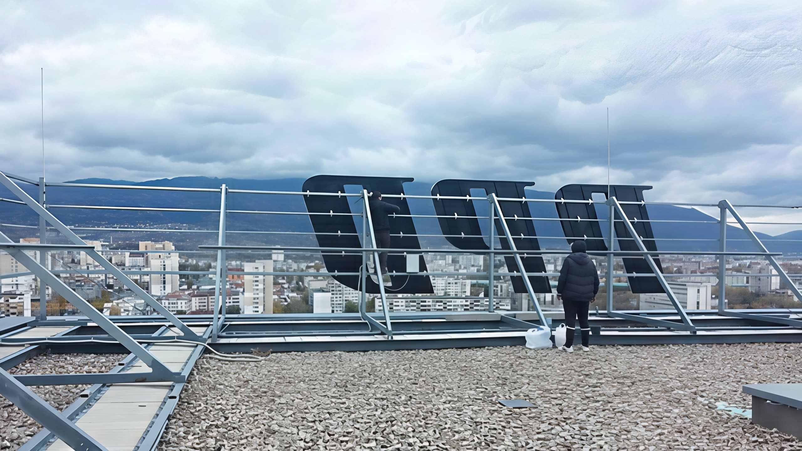 Работник до гърба на големи букви, монтирани на метална конструкция върху покрив с изглед към града.