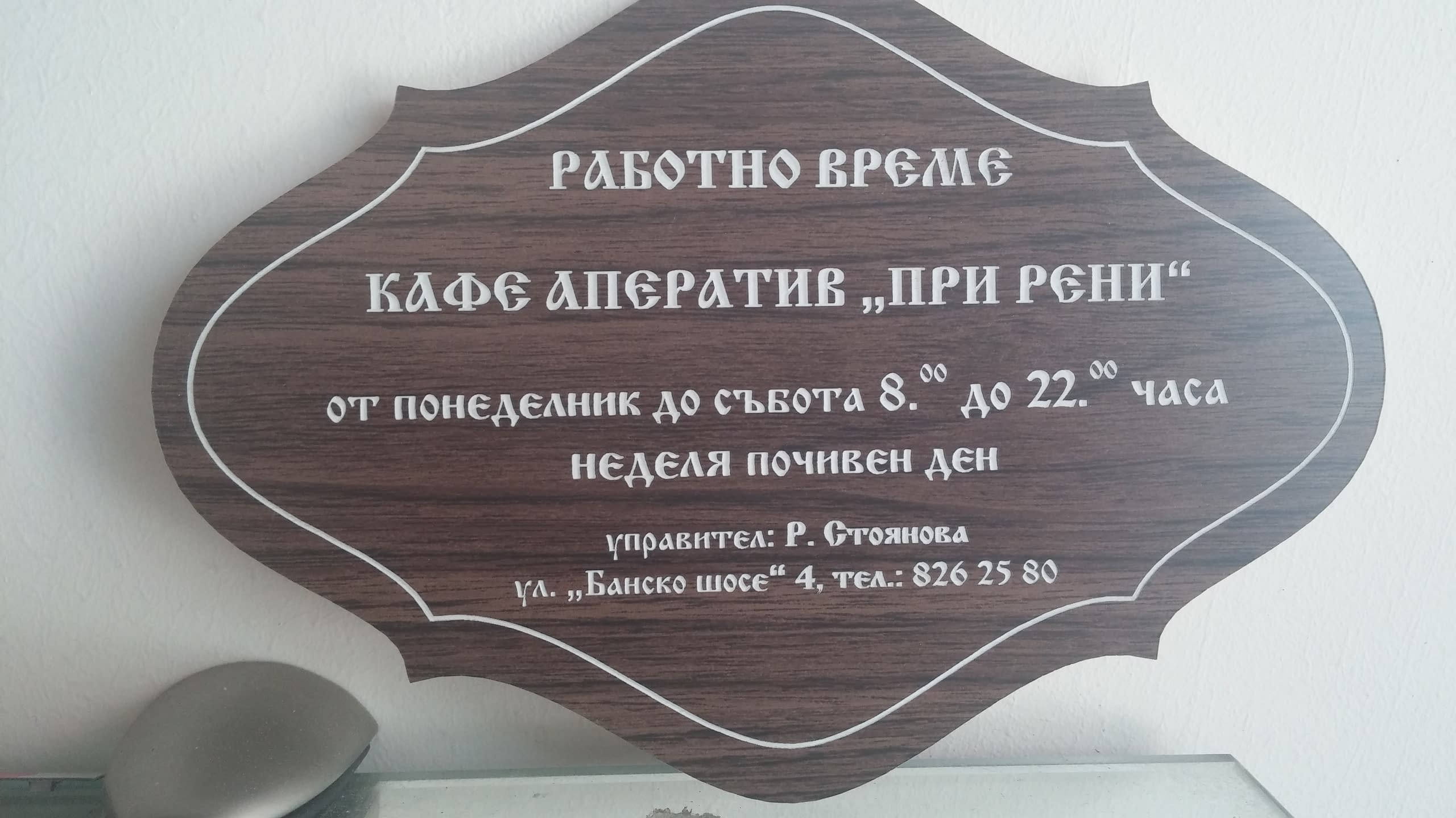 Табела с работно време за "Кафе Аператив При Рени" с фигурална форма и текстура на тъмно дърво.