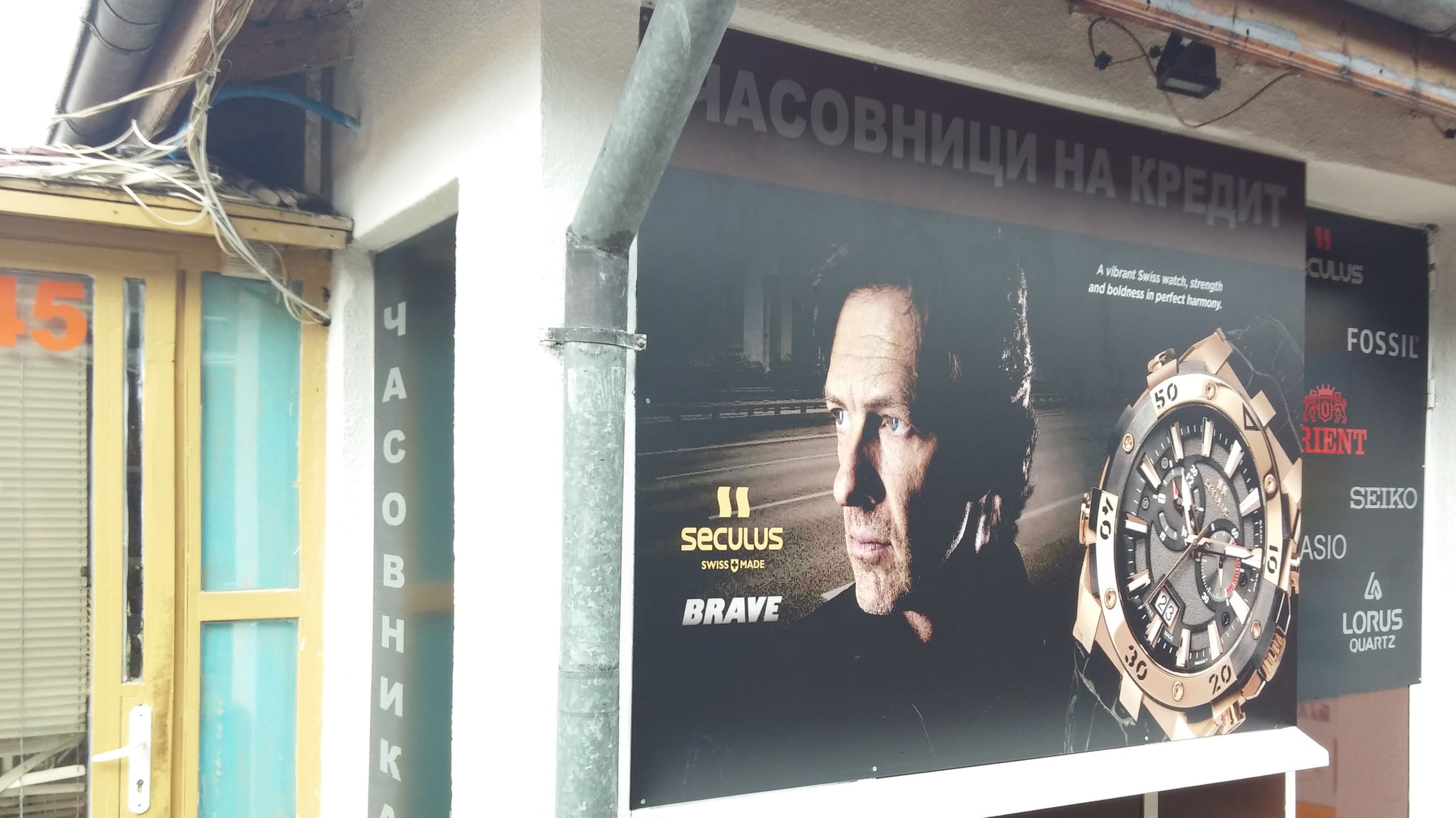 Страничен изглед на табела на часовникар, включваща вертикален надпис и рекламно изображение на часовник.