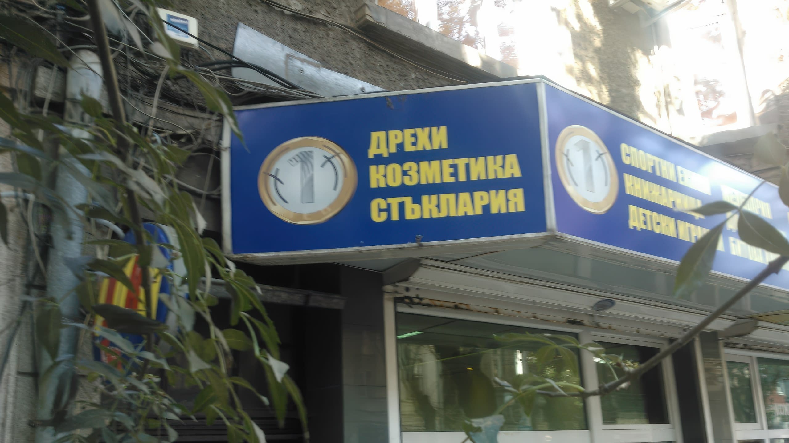 Ъглова светеща табела на магазин със син фон и жълти надписи за дрехи, козметика и стъклария.