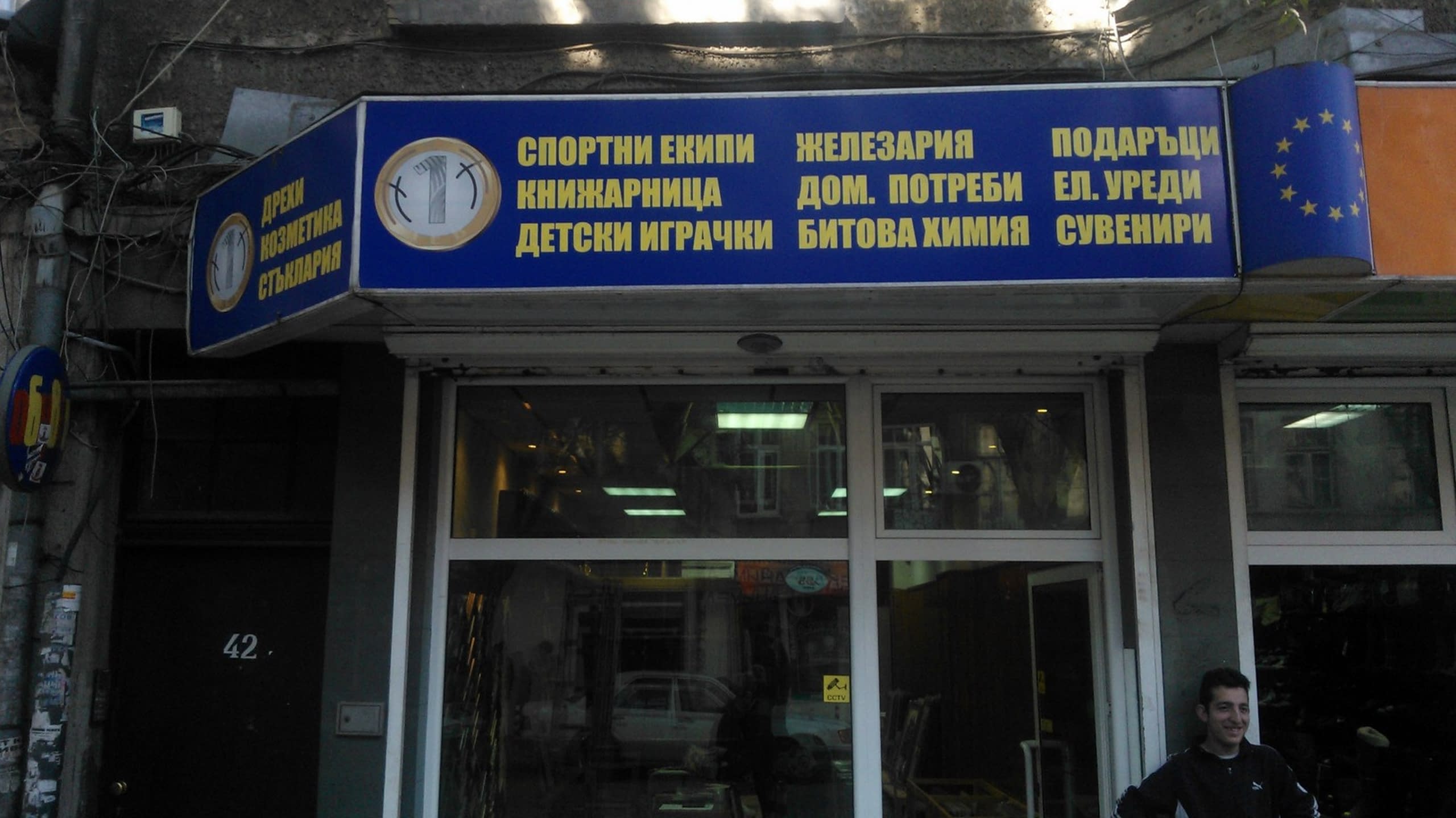 Синя светеща табела на магазин, изброяваща множество продуктови категории като книжарница и железария.