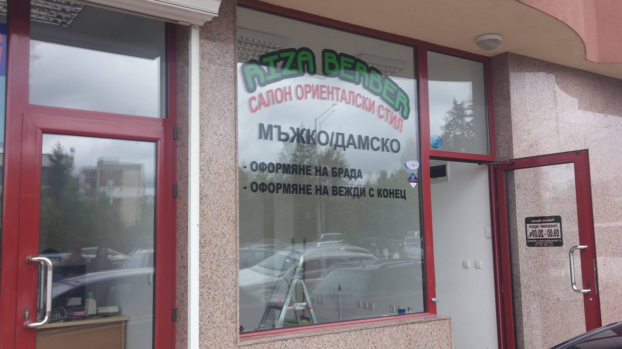 Стъклена витрина на фризьорски салон, облепена с надписи, погледната отстрани.