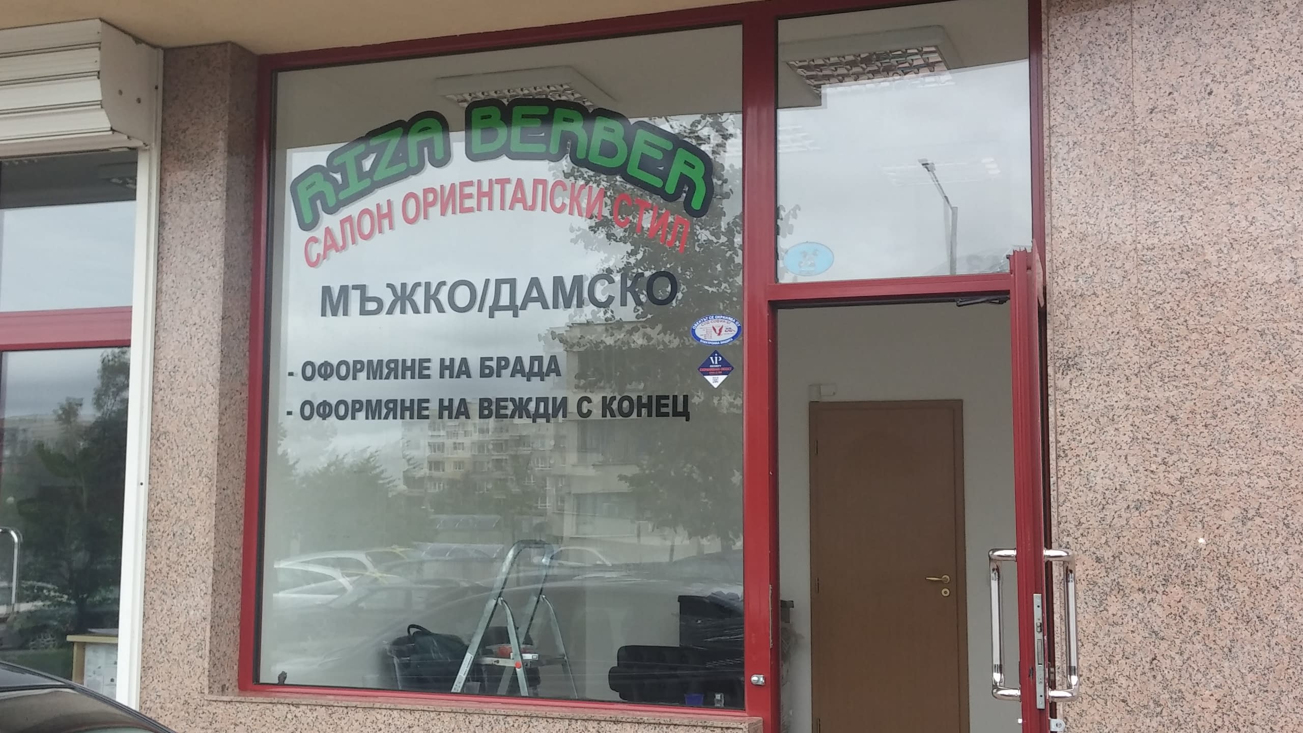 Надписи, залепени на прозореца на бръснарница, които описват услуги като оформяне на брада и вежди.