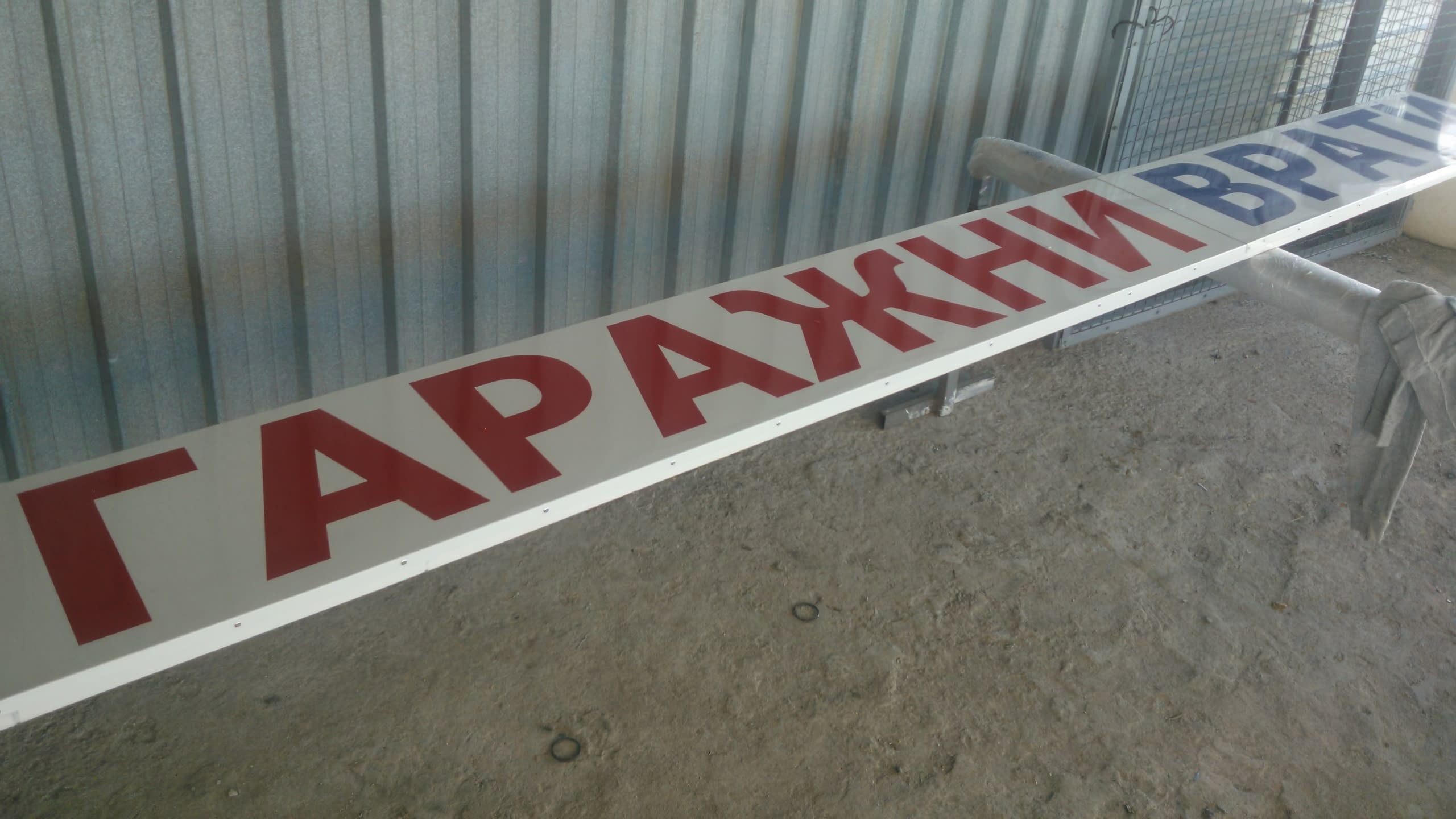 Изглед в близък план на лявата част на бяла табела, показващ червения надпис „ГАРАЖНИ“ на фона на ламаринена стена.