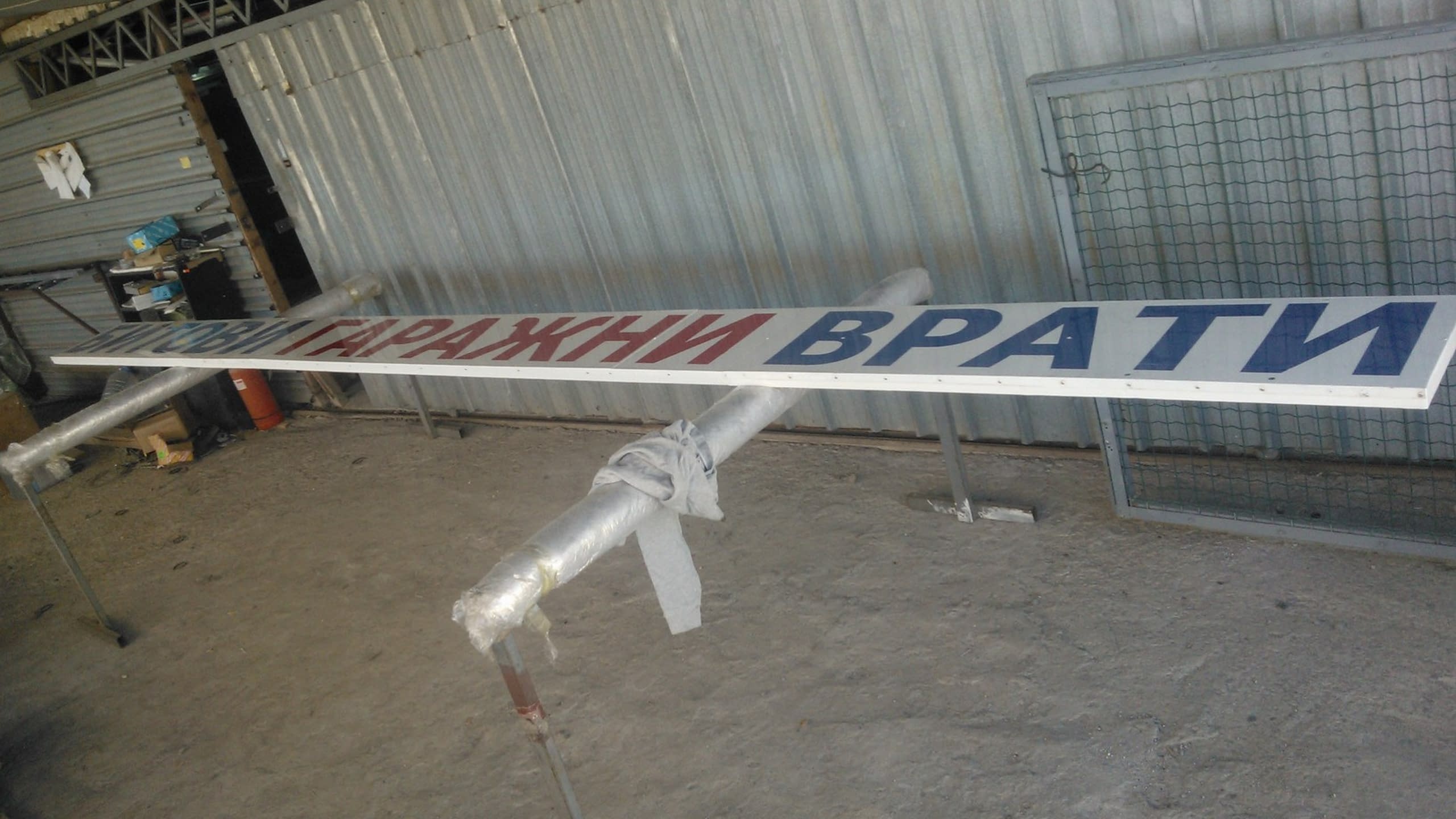 Дълга, правоъгълна табела с червен и син надпис на кирилица, подпряна на метални стойки в работно помещение с ламаринени стени.