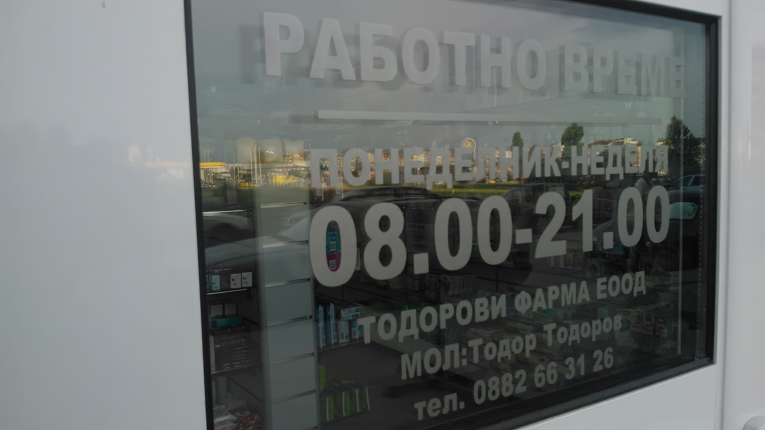 Изглед под ъгъл към стъклена витрина с бял надпис за работно време, в който се отразява външна среда и паркиран автомобил.