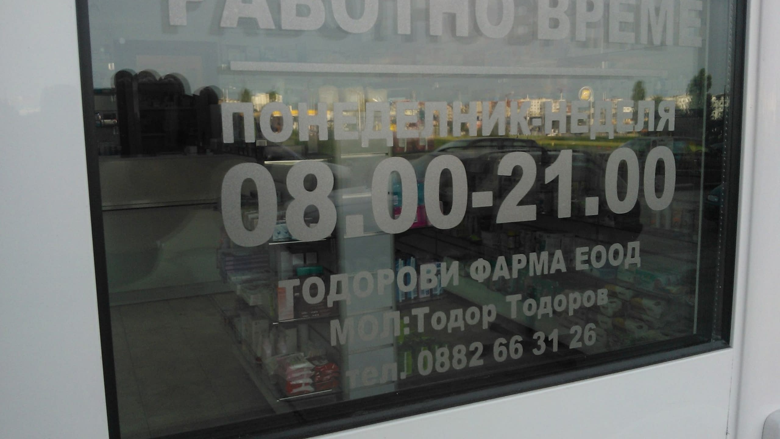 Стъклена врата на обект с апликиран бял текст на кирилица, указващ работно време от 08.00 до 21.00 и името на фирмата.
