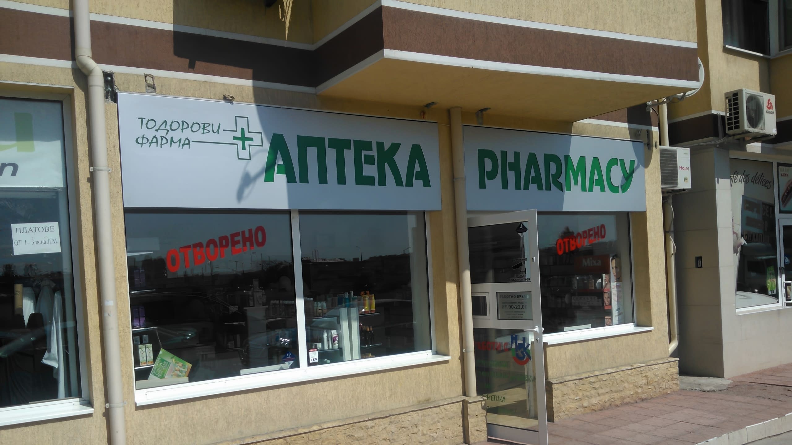 Външен изглед на ъглов магазин с две бели табели със зелени надписи на кирилица и латиница, разположени над витрините.