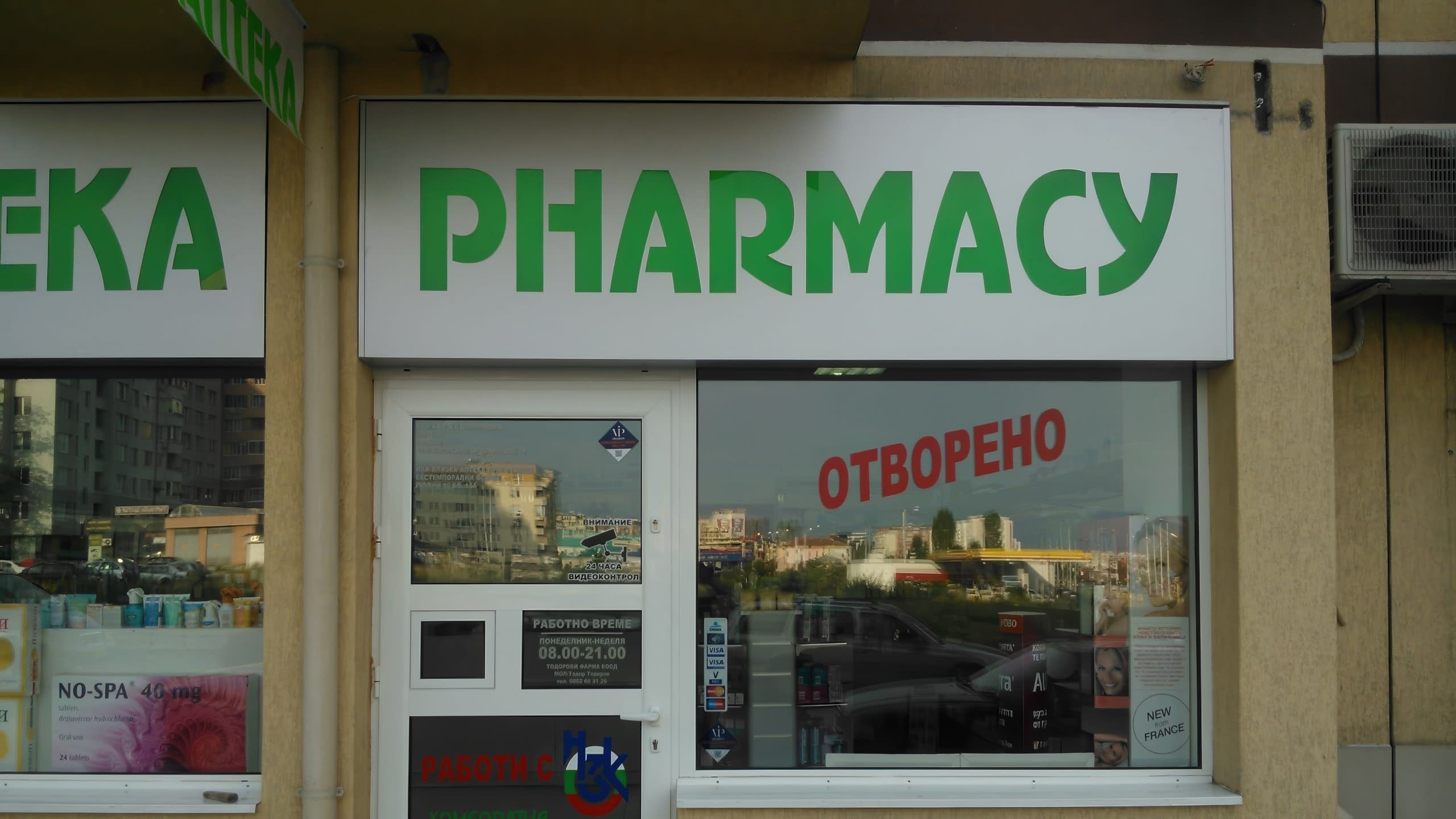 Вход на аптека, над който е поставена бяла табела със зелен надпис "PHARMACY", а на стъклената витрина има червен стикер.