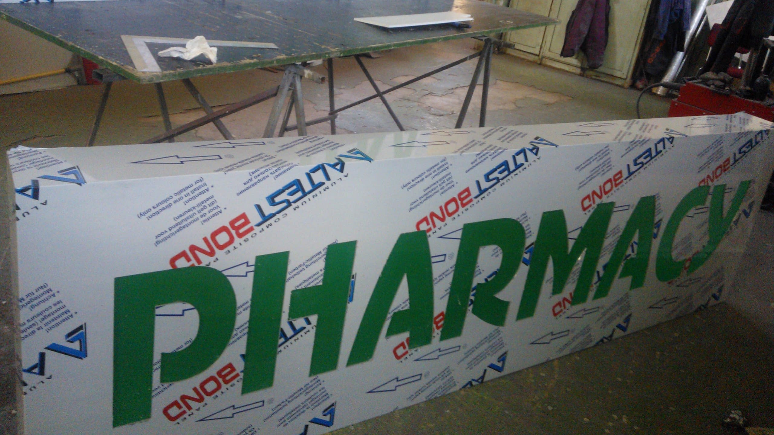 Голяма правоъгълна табела с изрязан зелен надпис "PHARMACY", покрита със защитно фолио с лога, заснета в работно помещение.